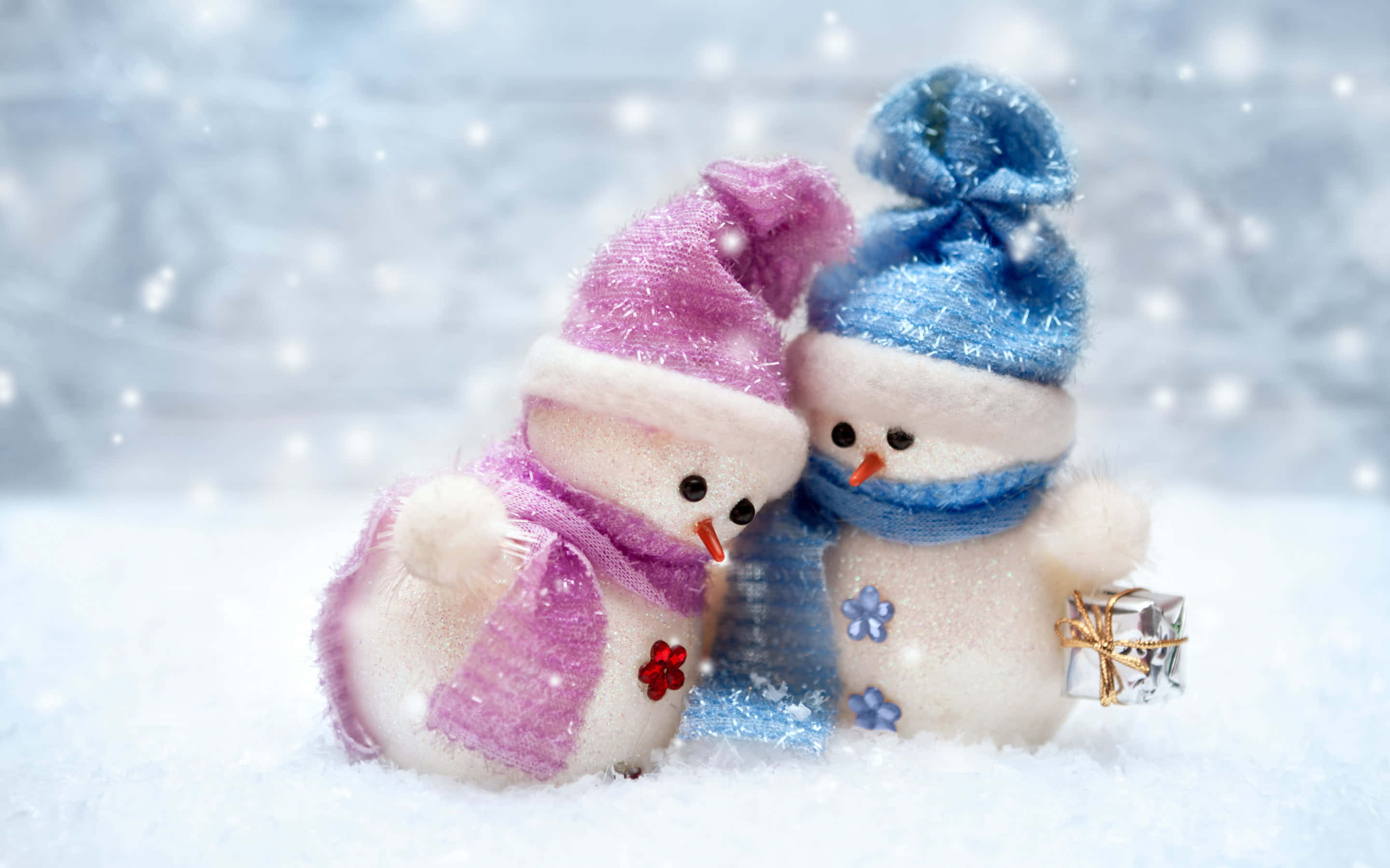 Couples Christmas 桌布 壁紙 — 兩個毛茸茸的雪人戴著色彩繽紛的帽子和圍巾，在飄雪中依偎，旁邊有一份禮物。