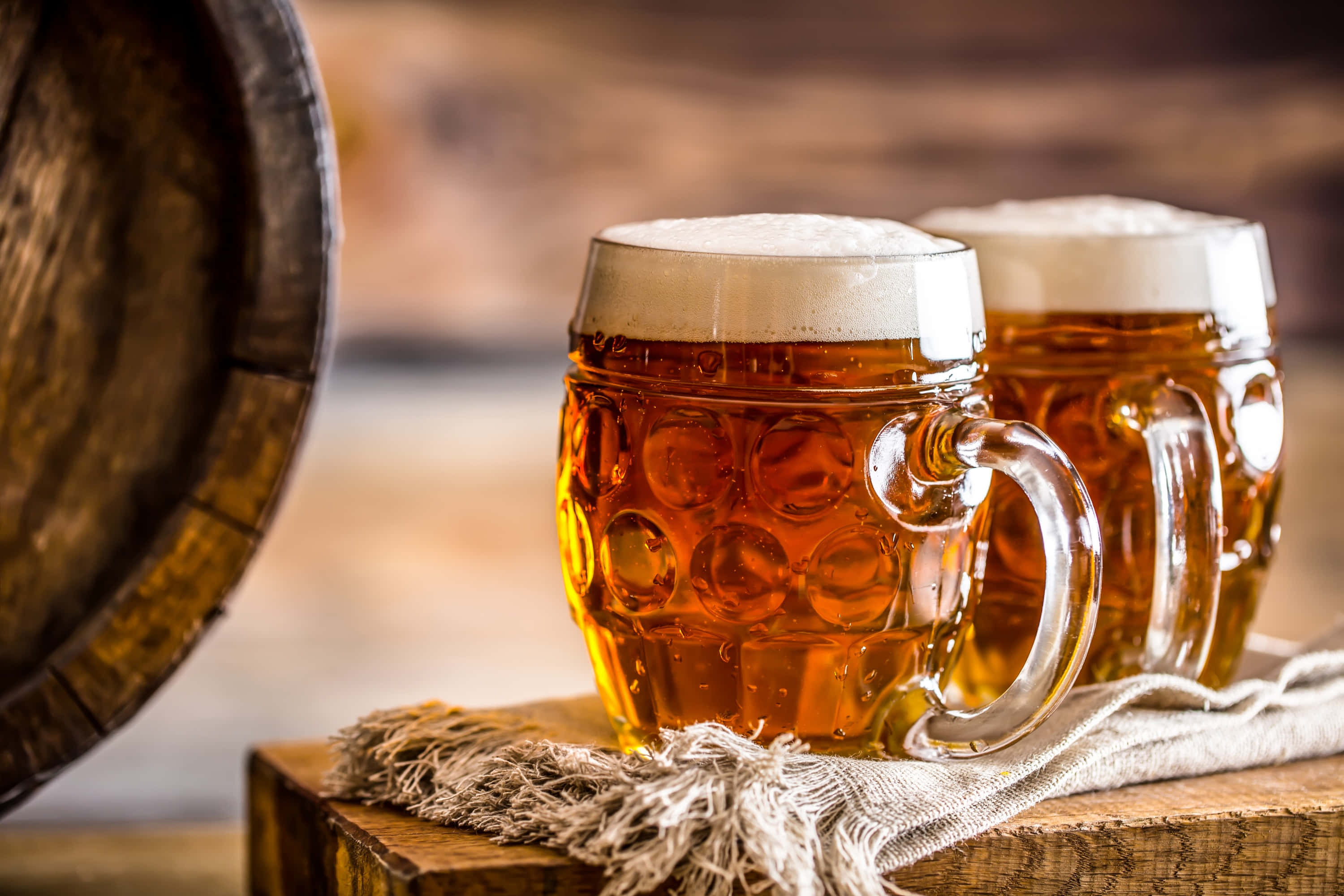 Beer 桌布 壁紙 — 兩個泡沫豐富的琥珀色啤酒杯，帶有氣泡紋理玻璃，放在一個質樸的木板上，旁邊有一個木桶。