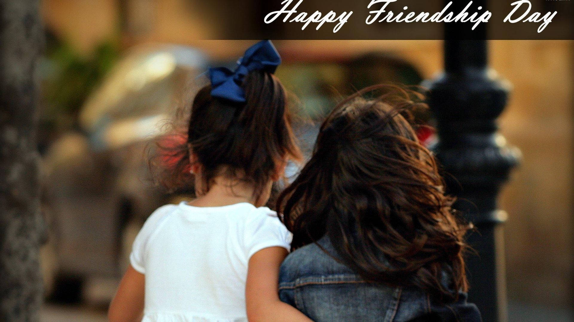 Friendship Day 桌布 壁紙 — 兩個深色頭髮、戴藍色蝴蝶結的年輕女孩手挽手走在陽光明媚的街道上。