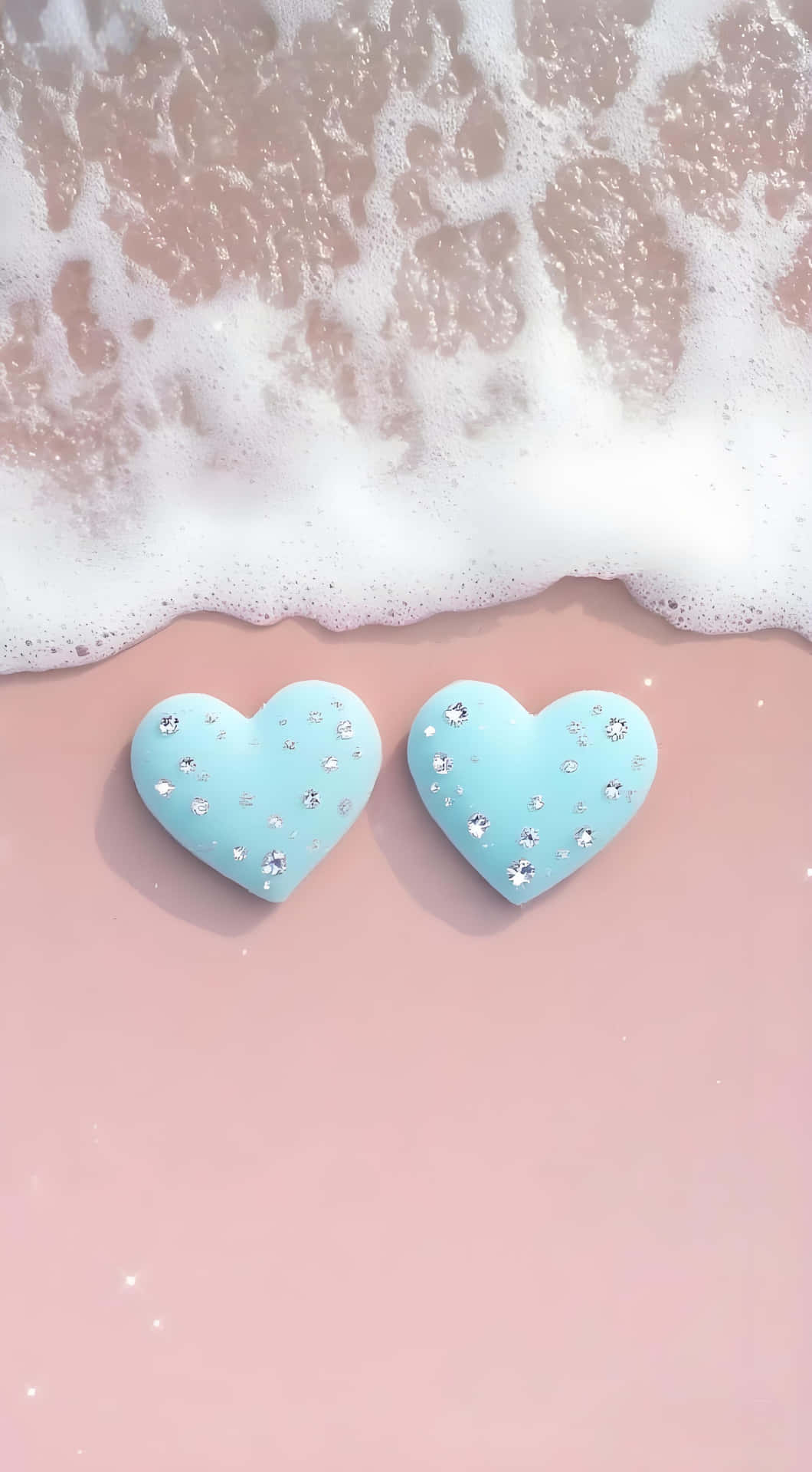 Pink & Blue Heart Diamonds: Fresh Romantic Wave Wallpaper 壁紙 — 兩個淺藍色心形物體，飾有閃亮水鑽，放在柔軟的粉紅色沙子上，溫柔的海浪泡沫湧入。