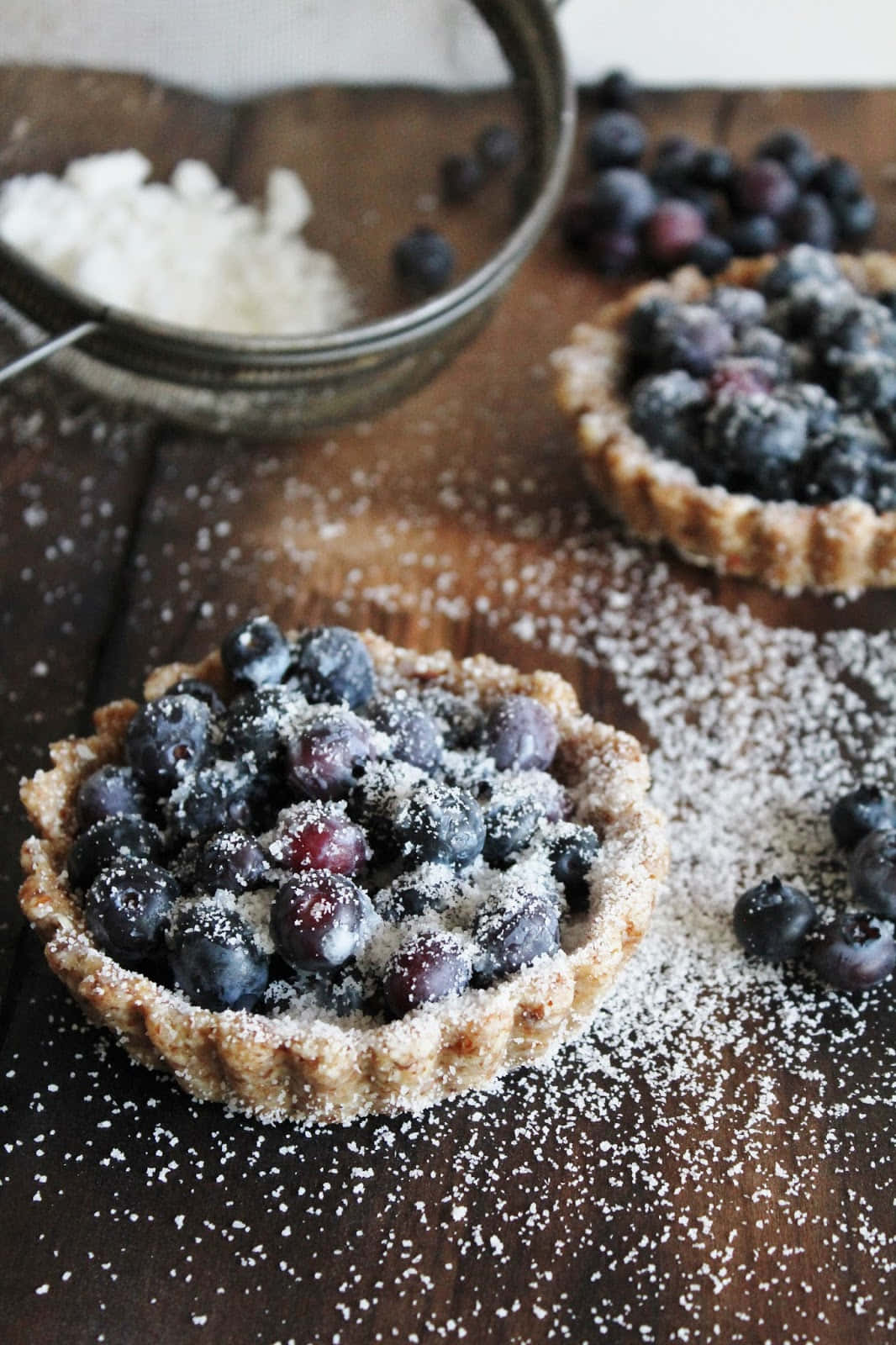 Blueberries Tart 桌布 壁紙 — 兩個灑滿糖霜的鄉村藍莓塔放在深色木桌上，旁邊有篩過的糖粉和新鮮莓果。