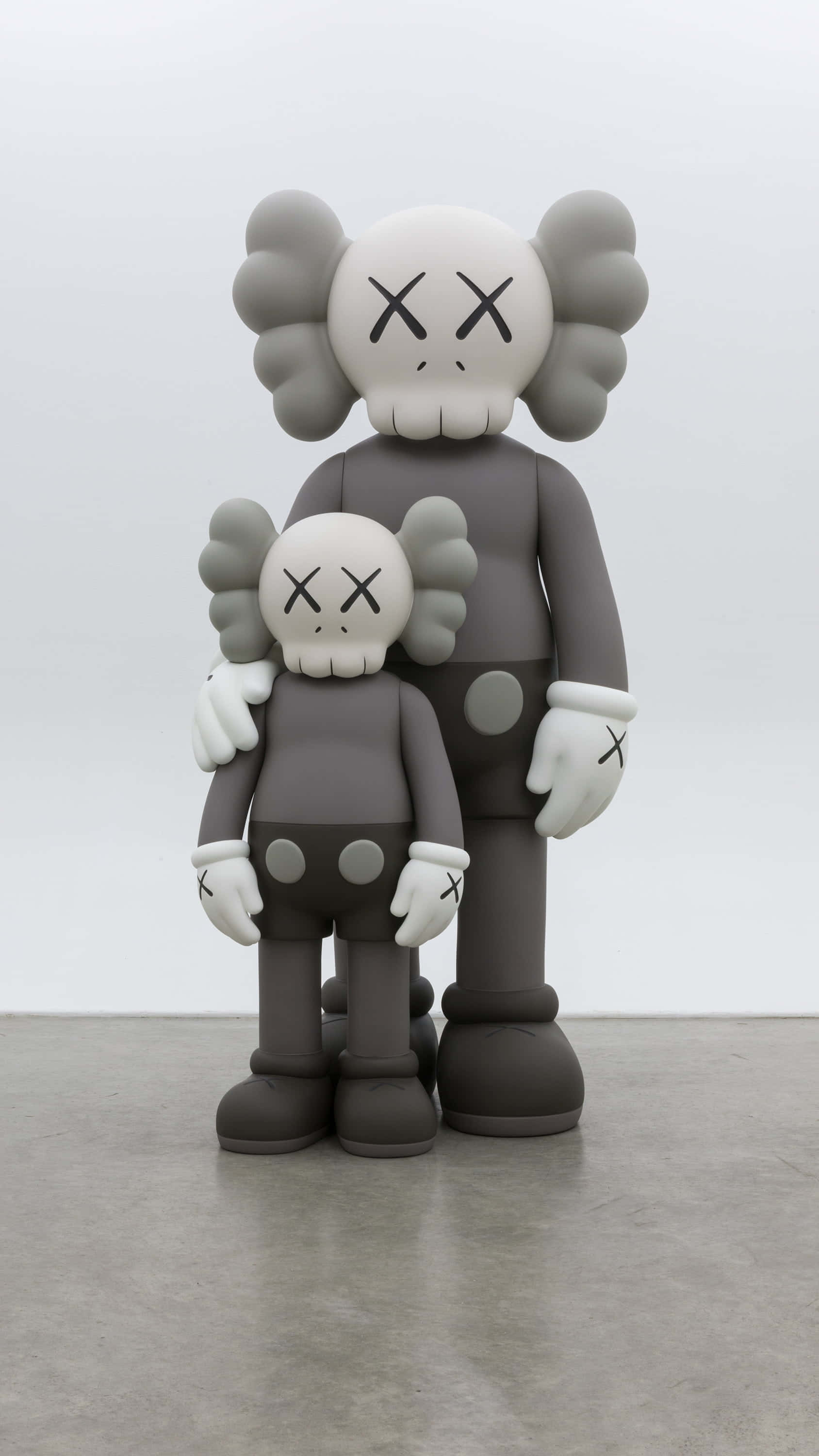 Kaws Bff 桌布 壁紙 — 兩個灰色KAWS Companion公仔，一大一小，並排站在一個簡約的畫廊空間，地板是水泥地，牆壁是白色的。