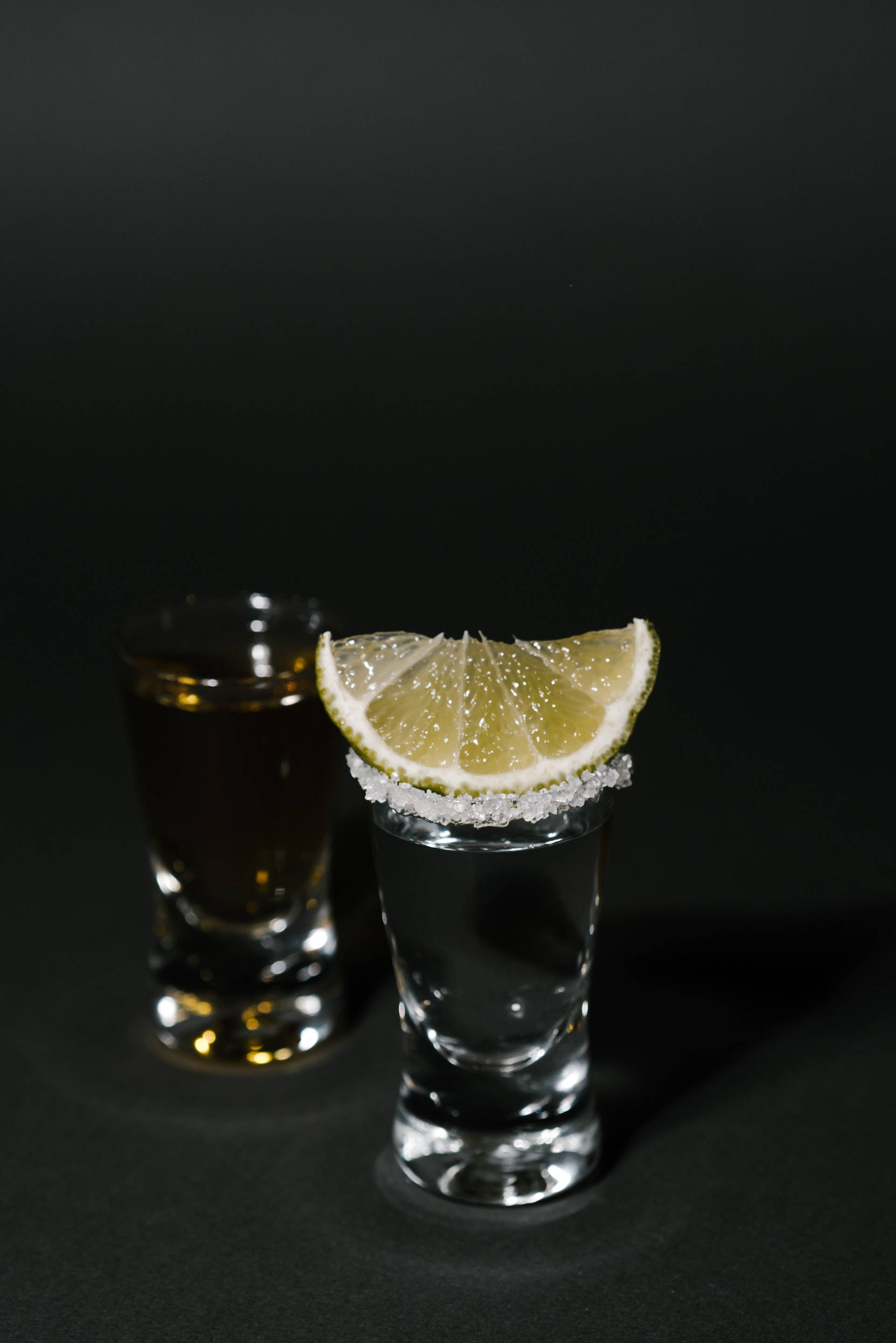 alcohol 桌布 壁紙 — 兩個烈酒杯，一個透明杯口沾鹽並飾以萊姆角，另一個深琥珀色，兩者均置於純黑背景前。