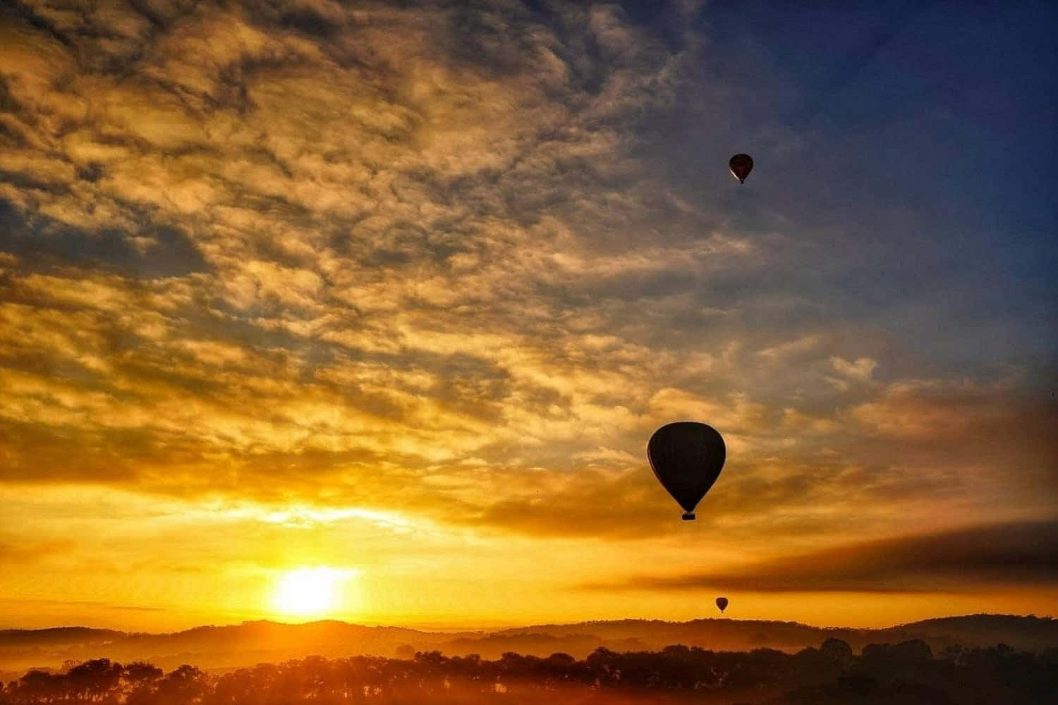 Hot Air Balloon 桌布 壁紙 — 兩個熱氣球在火紅的日落天空下，飄過起伏的山丘和樹木。