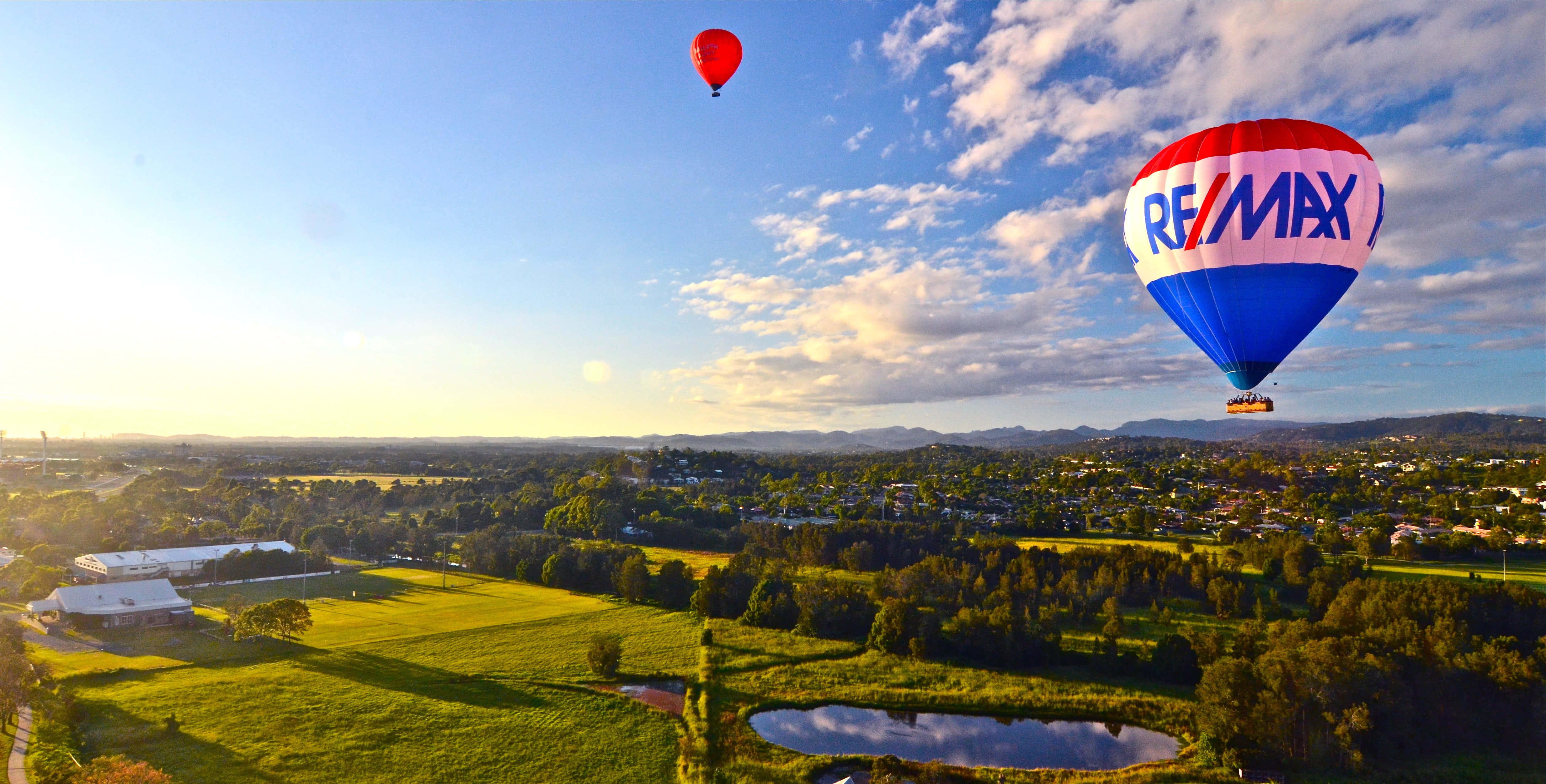 Hot Air Balloon 桌布 壁紙 — 兩個熱氣球在陽光明媚的山谷上空翱翔，山谷中有綠色田野和遠處的山丘，天空部分多雲。