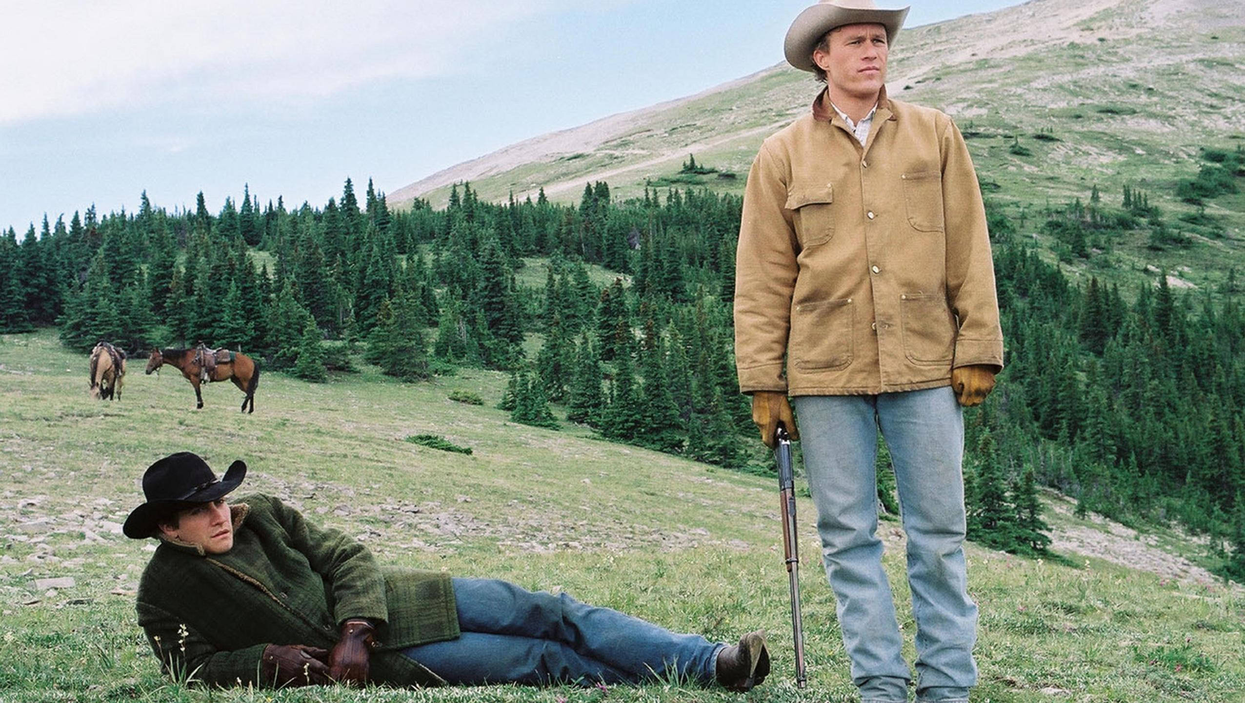 brokeback mountain 桌布 壁紙 — 兩個牛仔，一個站著拿步槍，另一個躺在草地上，後面有馬吃草、松樹和山丘。