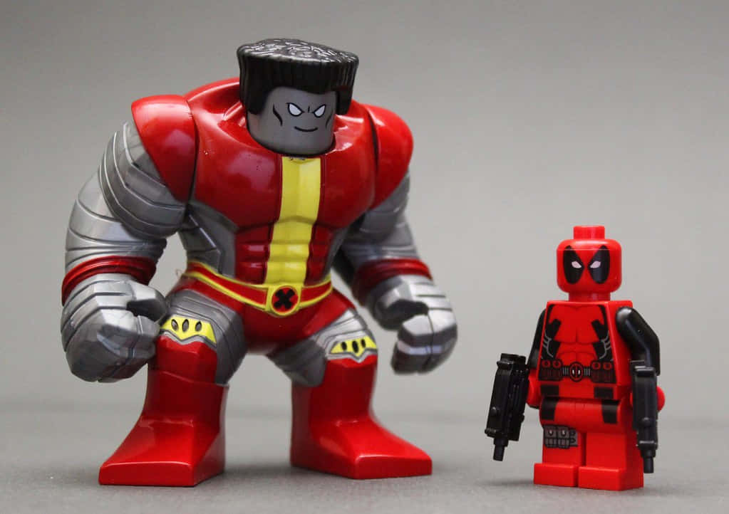Deadpool And Colossus 桌布 壁紙 — 兩個玩具動作人偶，一個大的紅色和銀色肌肉發達的人物和一個小的紅色死侍人偶，並排站在純灰色背景前。