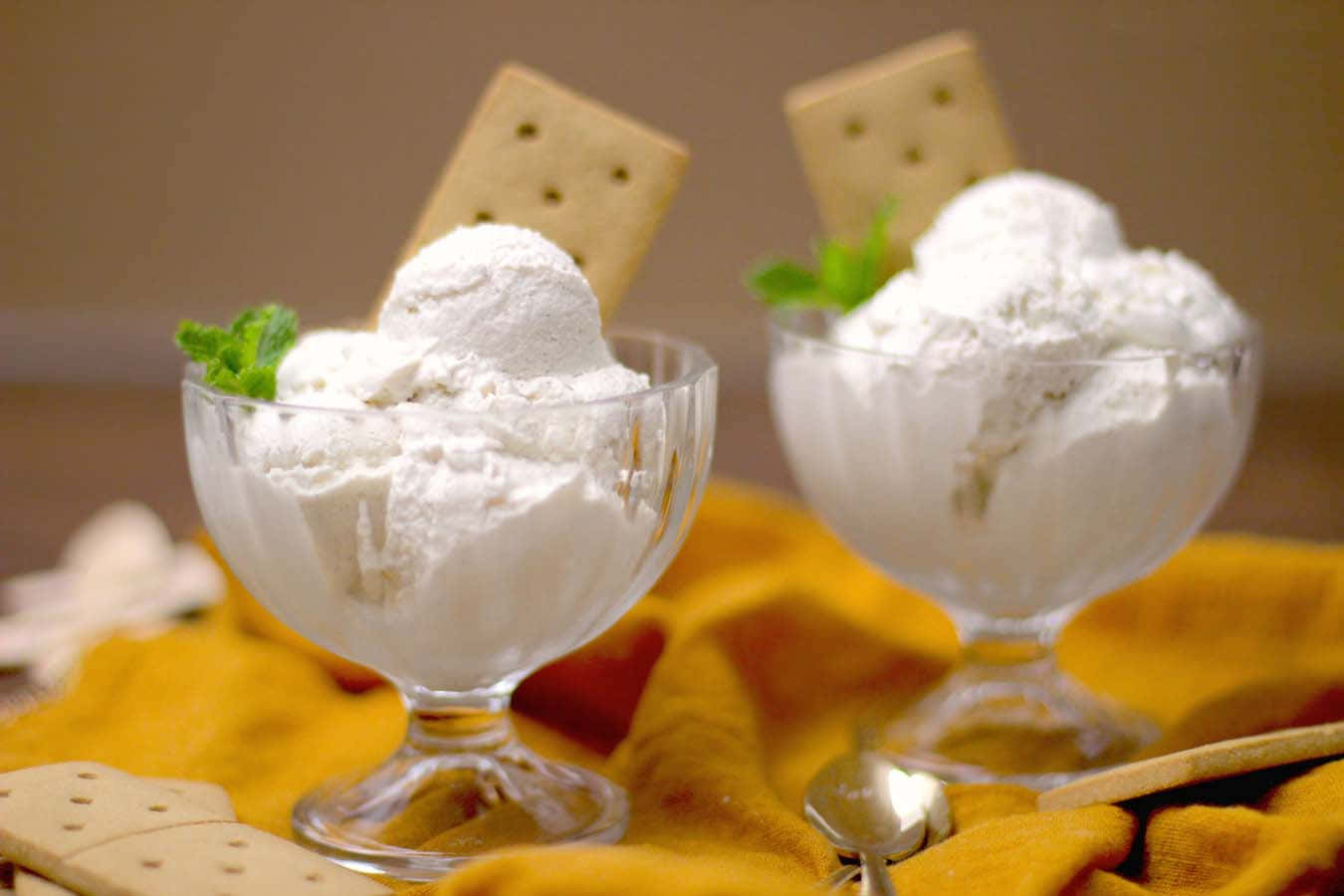 Ice Cream 桌布 壁紙 — 兩個玻璃碗裝滿奶油香草冰淇淋，上面撒上全麥餅乾和薄荷葉，放在溫暖的黃色布上。