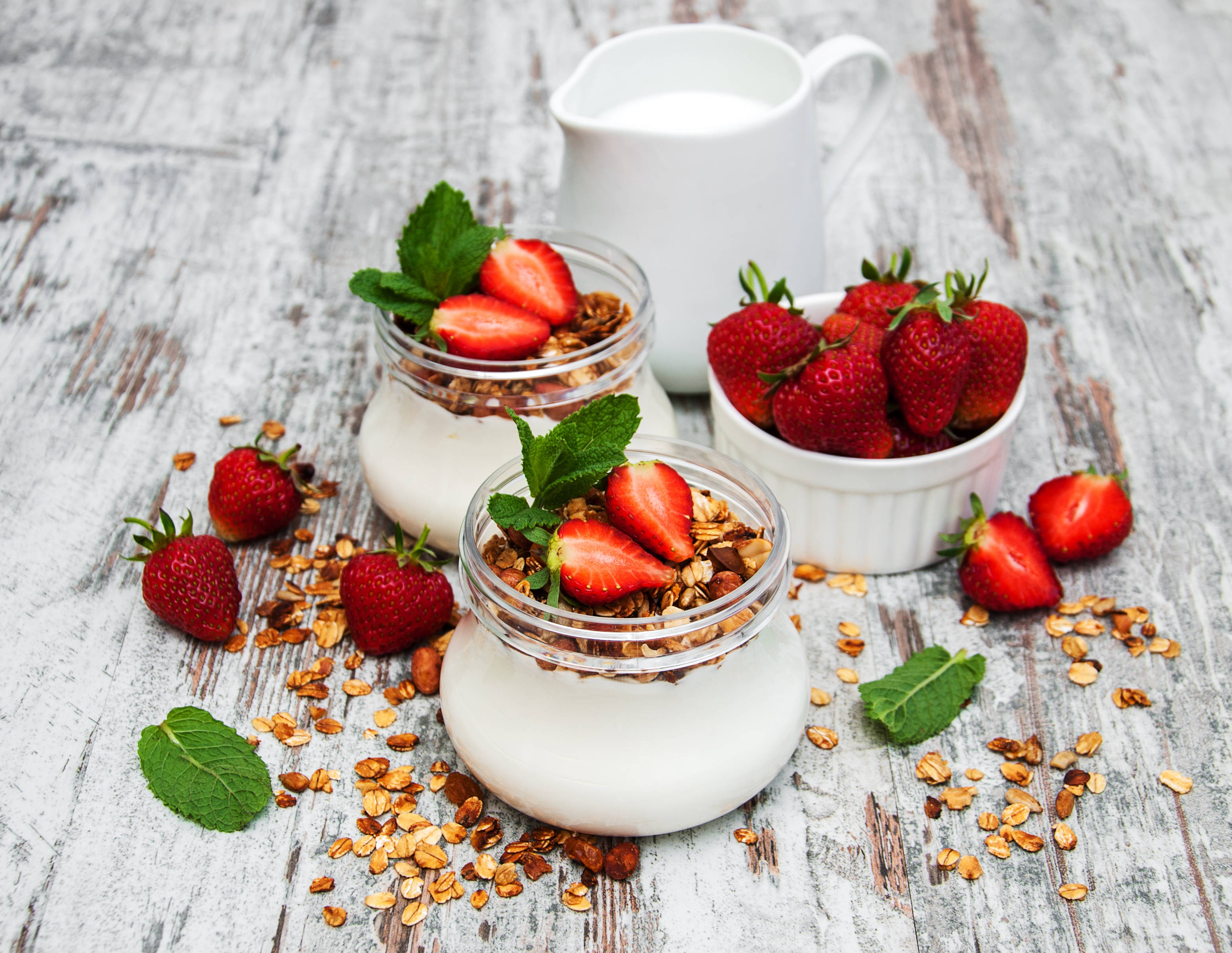 Yogurt 桌布 壁紙 — 兩個玻璃罐裝滿了奶油優格，上面撒上燕麥片和新鮮草莓，放在一個經過風化的白色木桌上。