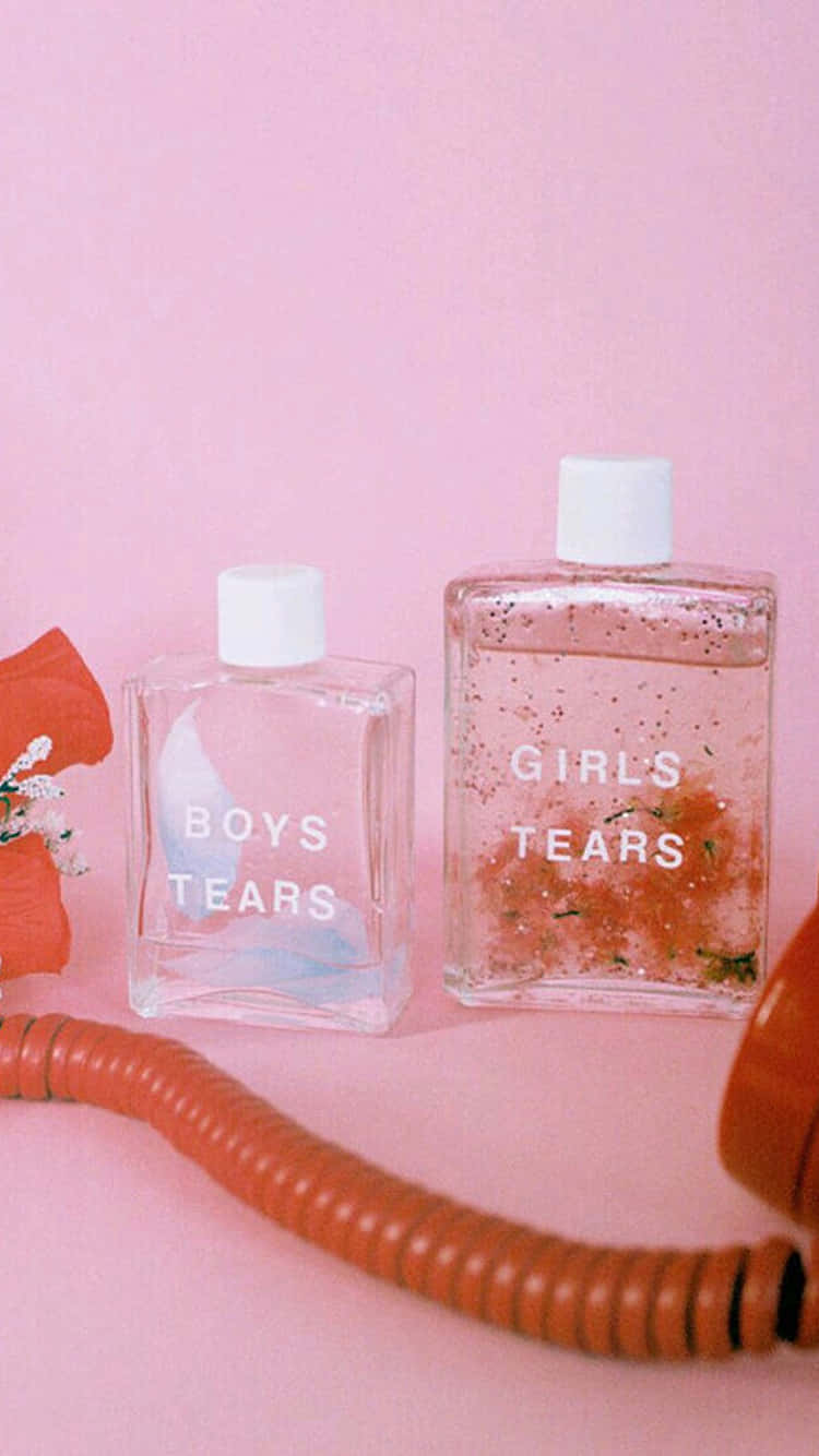 Pastel Vintage iPhone 桌布 壁紙 — 兩個玻璃香水瓶，標籤為「男孩的眼淚」和「女孩的眼淚」，放在粉紅色表面上，旁邊有一個復古橙色電話和一朵紅花。