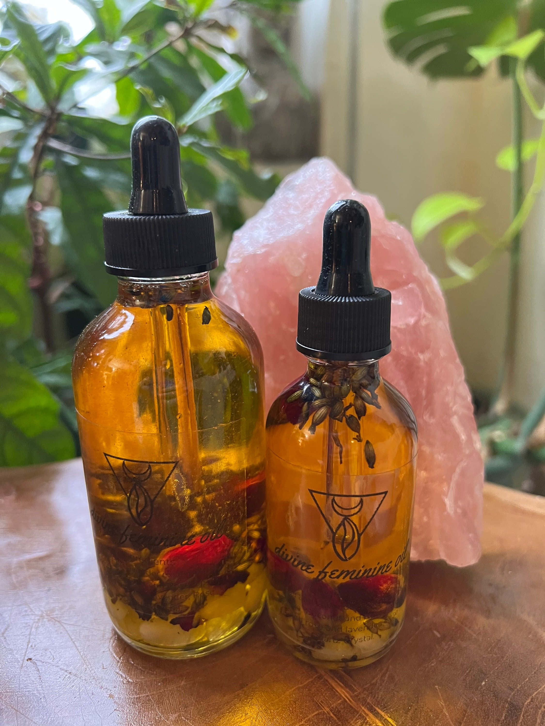 Rose Oil 桌布 壁紙 — 兩個琥珀色玻璃瓶裝滿草藥油和滴管，放在木桌上，旁邊有一個粉紅色水晶，周圍是茂盛的綠色植物。