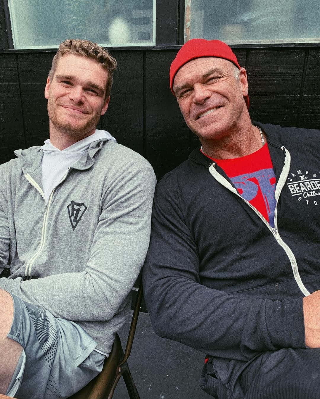 WWE 桌布 壁紙 — 兩個男人並排坐在深色木牆前，身後有窗戶，溫暖地微笑。