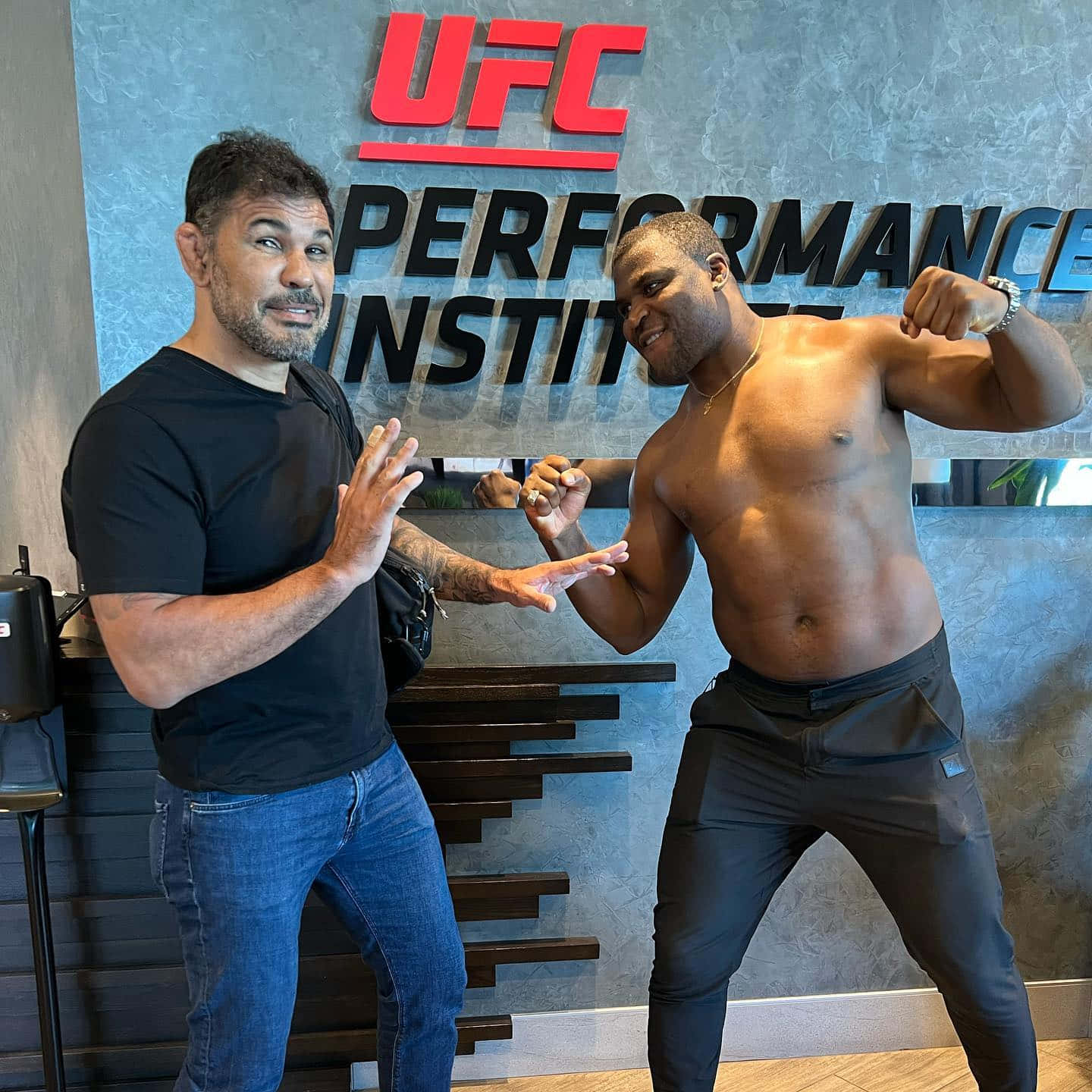UFC 桌布 壁紙 — 兩個男人在UFC Performance Institute標誌前嬉戲地擺姿勢，一個赤裸上身展示肌肉，另一個用手做手勢。