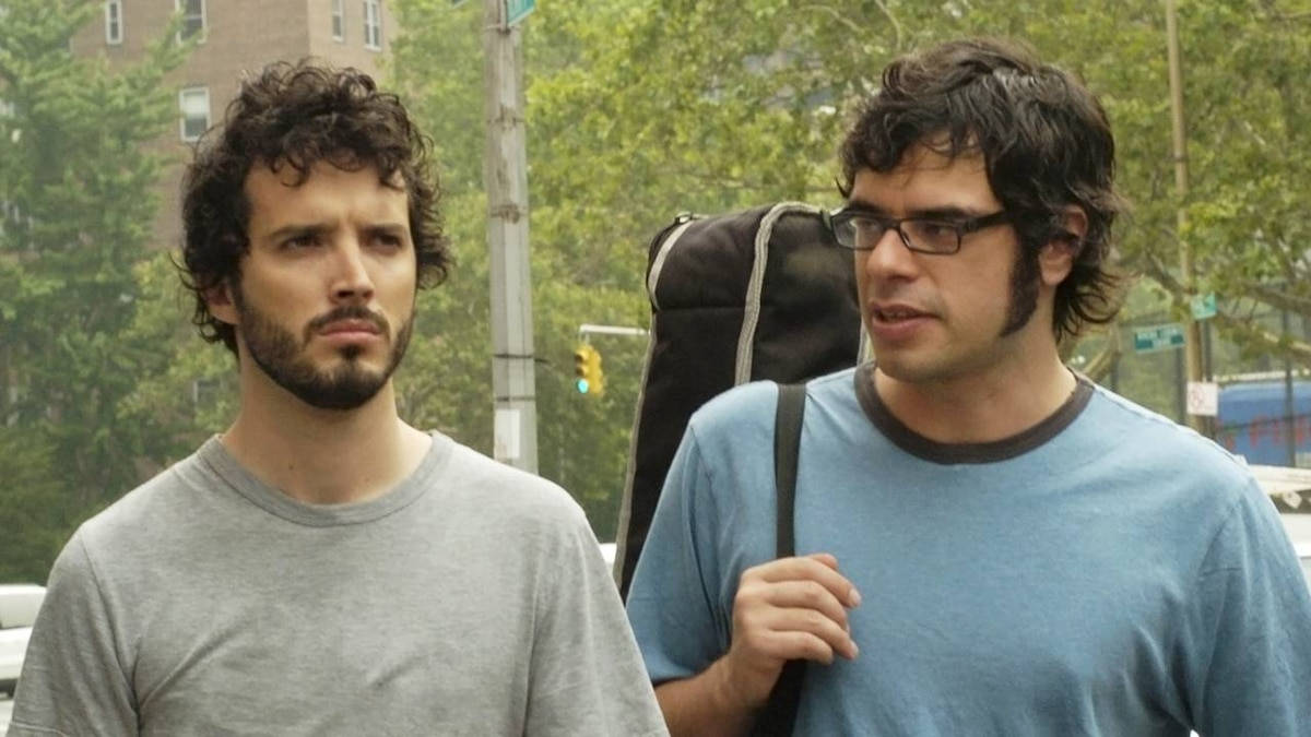 Flight Of The Conchords 桌布 壁紙 — 兩個男人在城市的街道上行走，其中一人拿著吉他盒，另一人則以嚴肅的表情向前看。