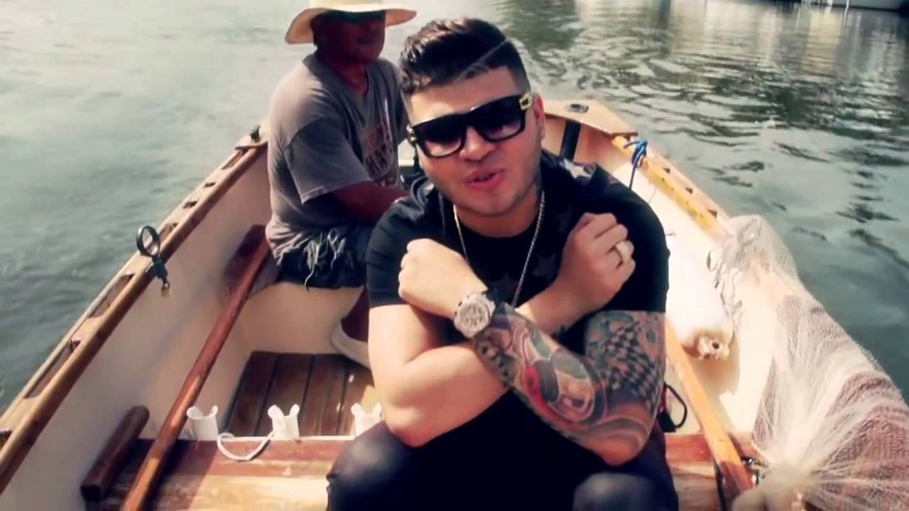 farruko 桌布 壁紙 — 兩個男人在平靜水面的木船上，一個有刺青和太陽眼鏡的人對著鏡頭擺姿勢，另一個在後面划船。