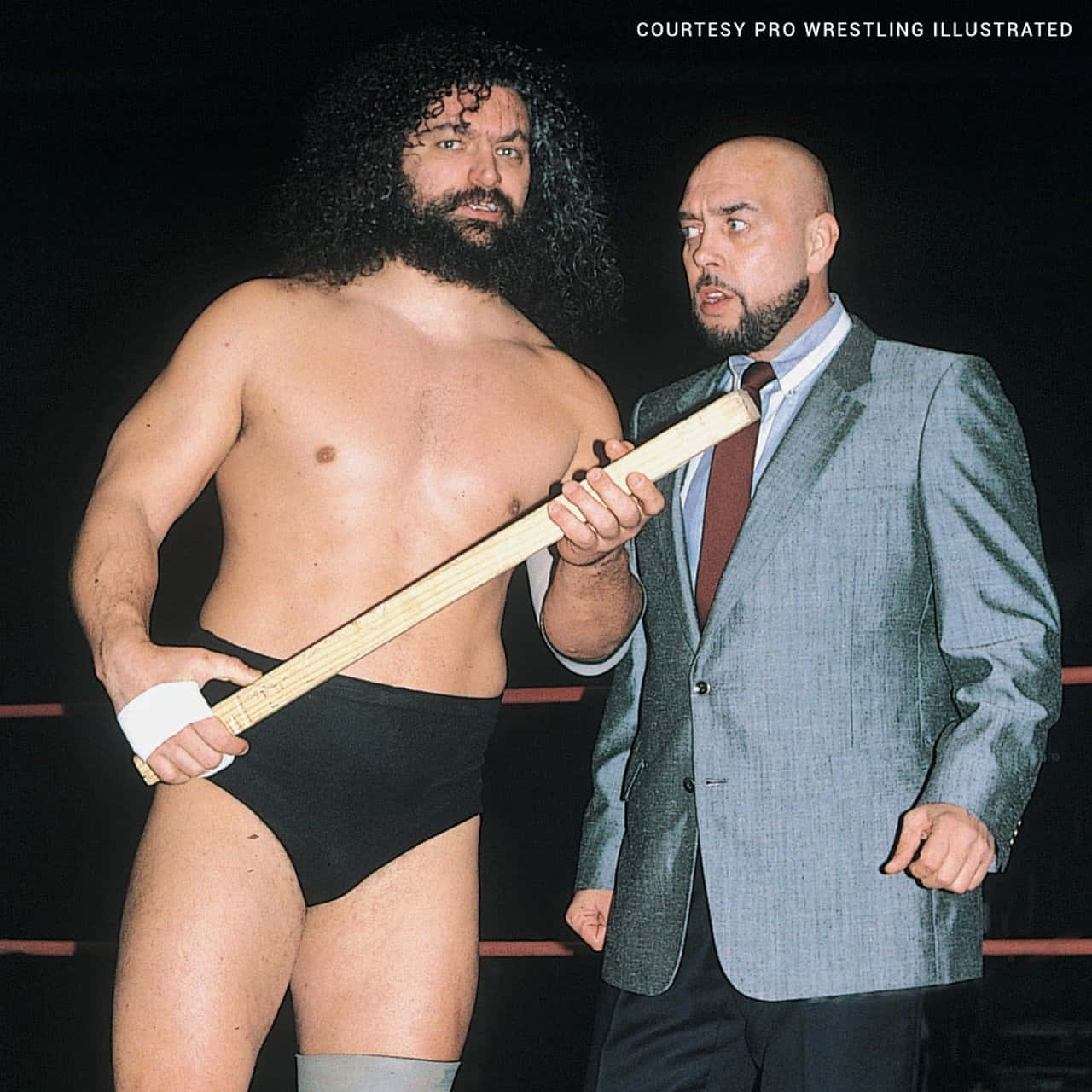 WWE 桌布 壁紙 — 兩個男人在摔角擂台上，一個赤裸上身拿著木棒，另一個穿著西裝，兩人都表情嚴肅。