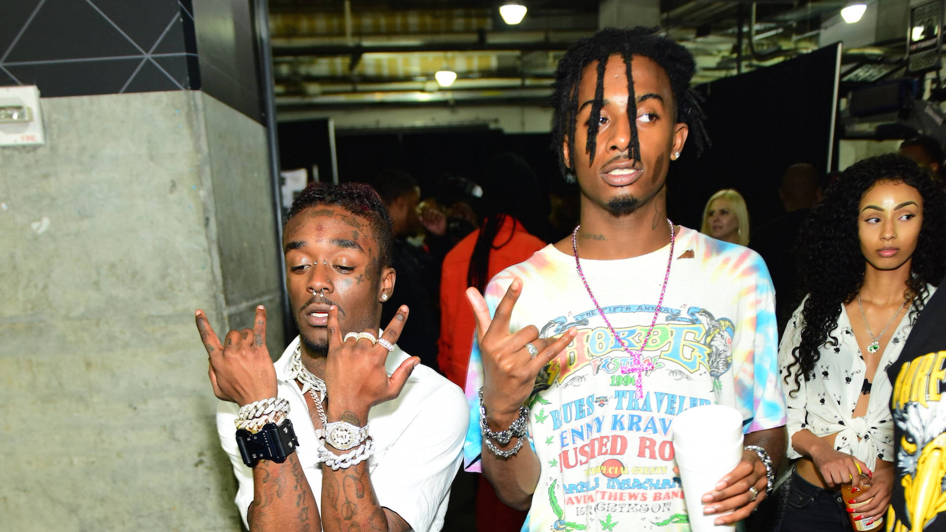 Carti 桌布 壁紙 — 兩個男人在擁擠的室內空間做出搖滾手勢，戴著閃亮珠寶和紮染襯衫。