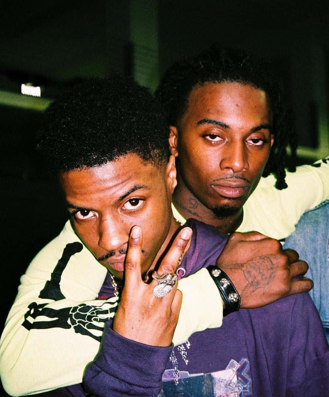 Playboi Carti 桌布 壁紙 — 兩個男人在昏暗燈光下親密擺姿勢，一個做手勢，另一個將手臂搭在他肩上。