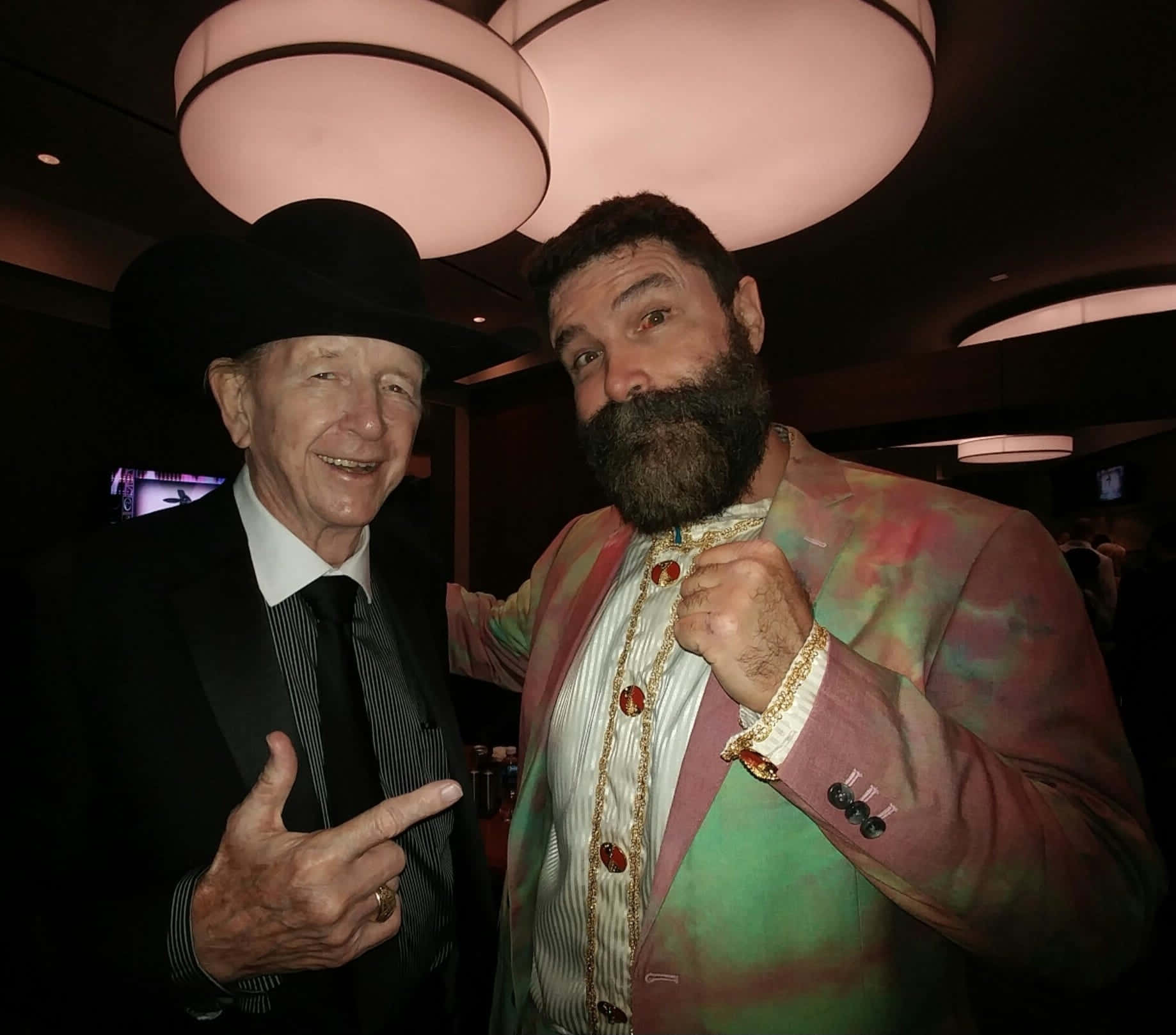 WWE 桌布 壁紙 — 兩個男人在昏暗的房間裡一起擺姿勢，一個穿著黑色西裝和帽子，另一個穿著彩色圖案外套，留著鬍子。