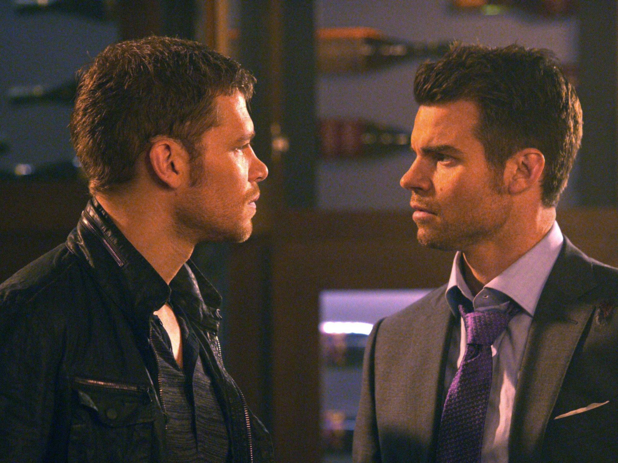 elijah mikaelson 桌布 壁紙 — 兩個男人在昏暗的房間裡對峙，表情緊張，衣著對比鮮明，一個穿皮衣，另一個穿西裝。