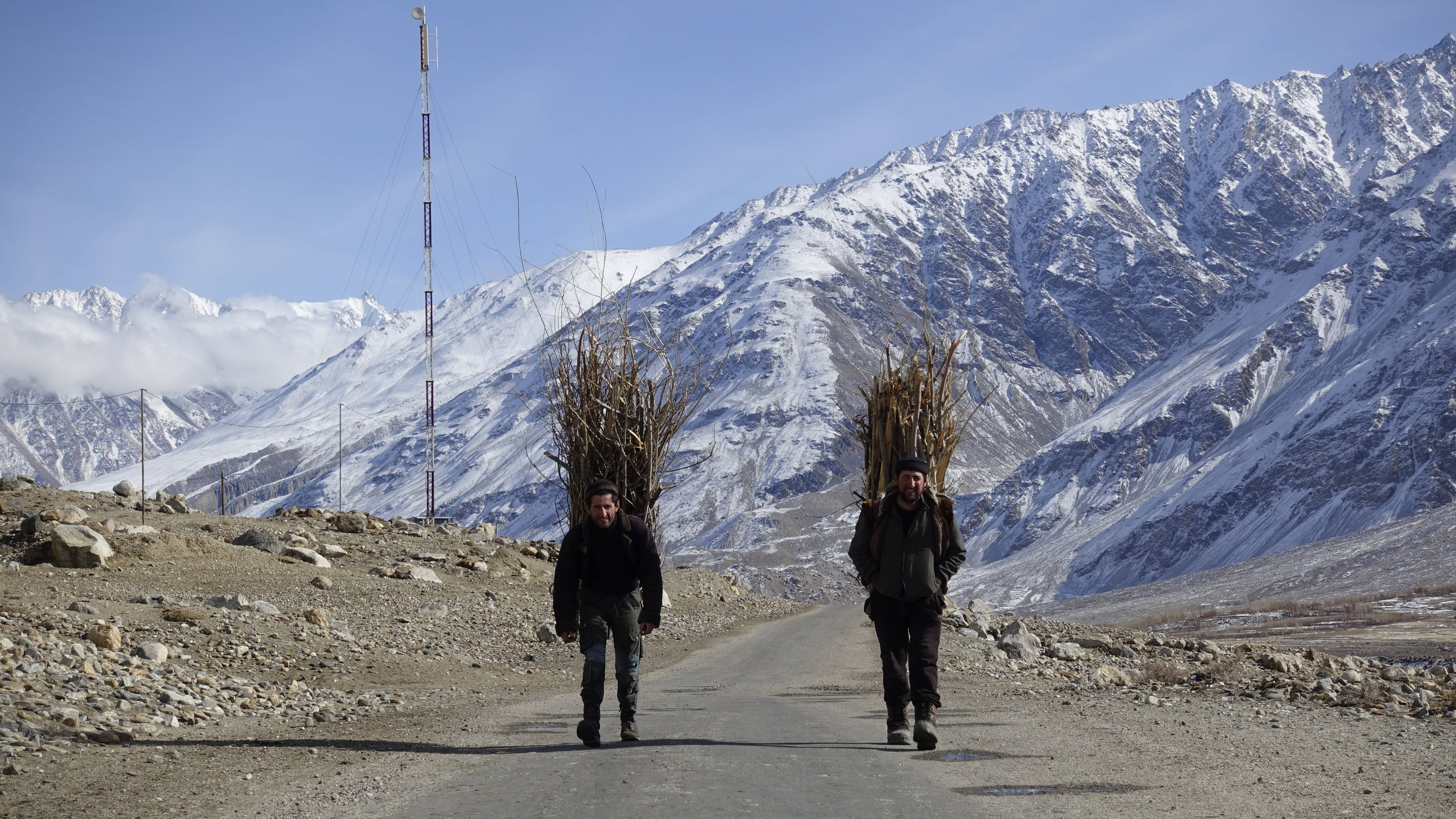 Tajikistan 桌布 壁紙 — 兩個男人在晴朗的藍天下，以高聳的雪山為背景，沿著碎石路扛著沉重的乾樹枝捆。