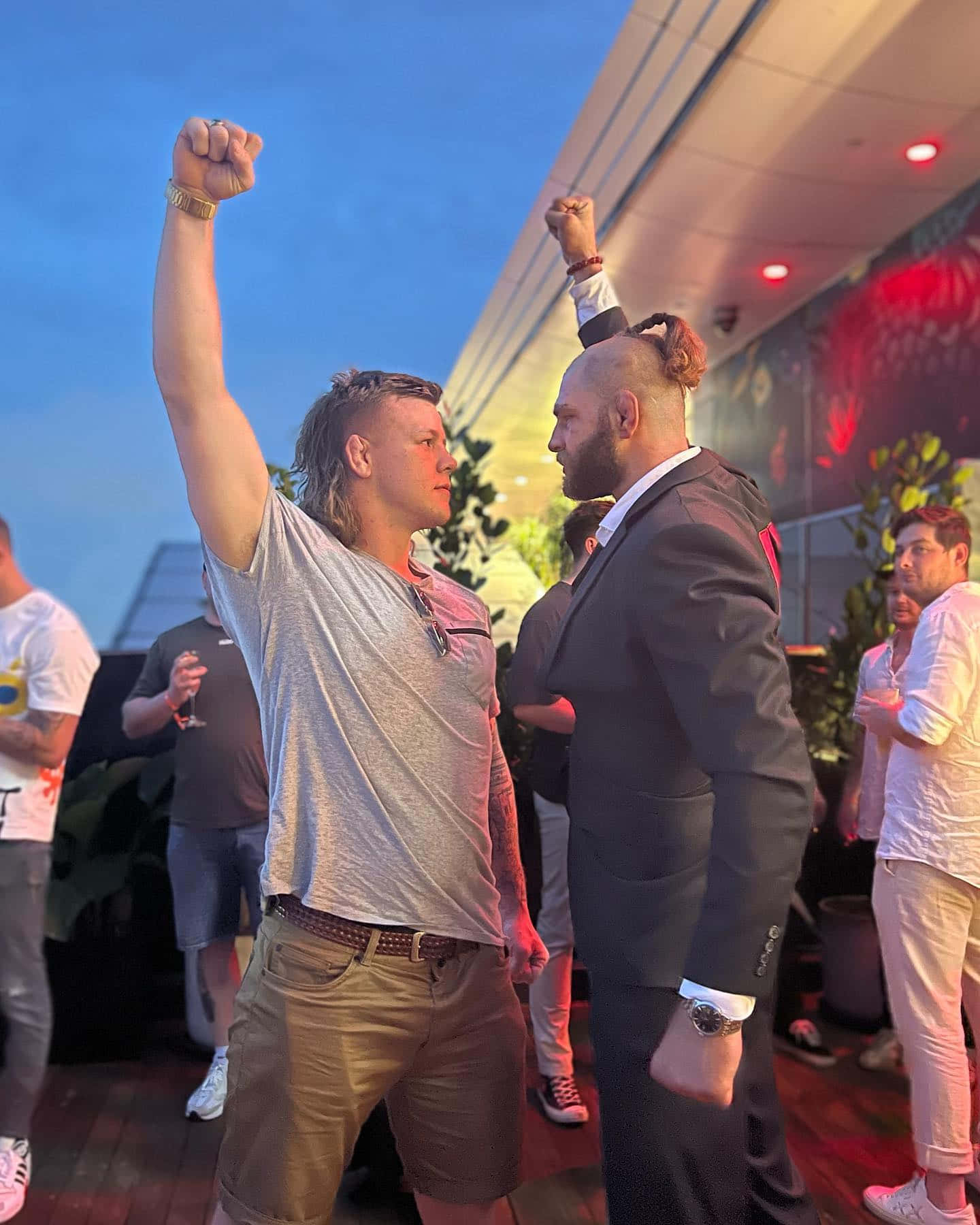 Cheering 桌布 壁紙 — 兩個男人在暮色天空下的屋頂派對中高舉拳頭慶祝勝利，周圍有閃爍的燈光和賓客。
