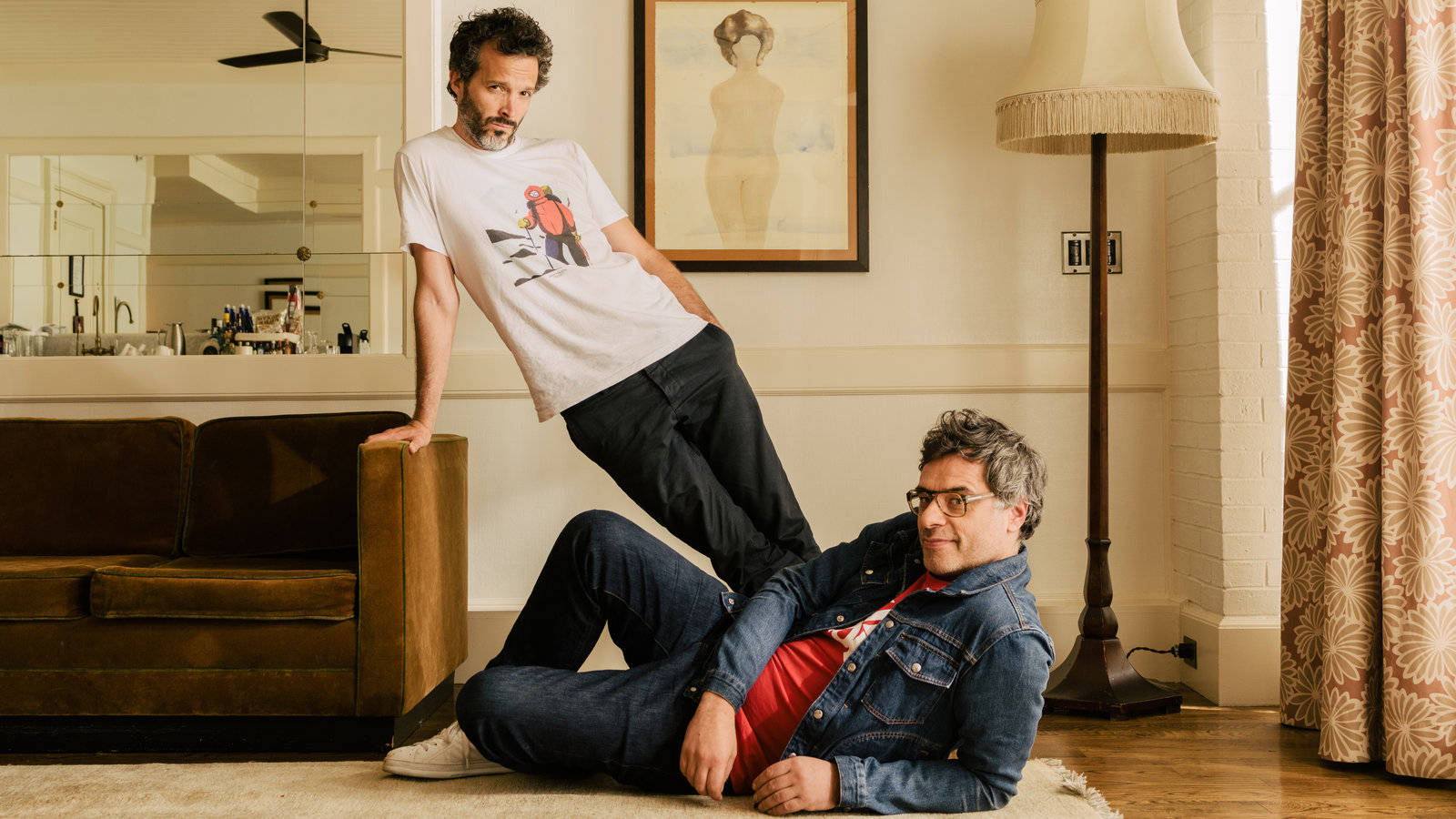 Flight Of The Conchords 桌布 壁紙 — 兩個男人在溫暖照明的客廳中，一個穿著牛仔外套躺在地板上，另一個穿著圖案T恤靠在沙發上。