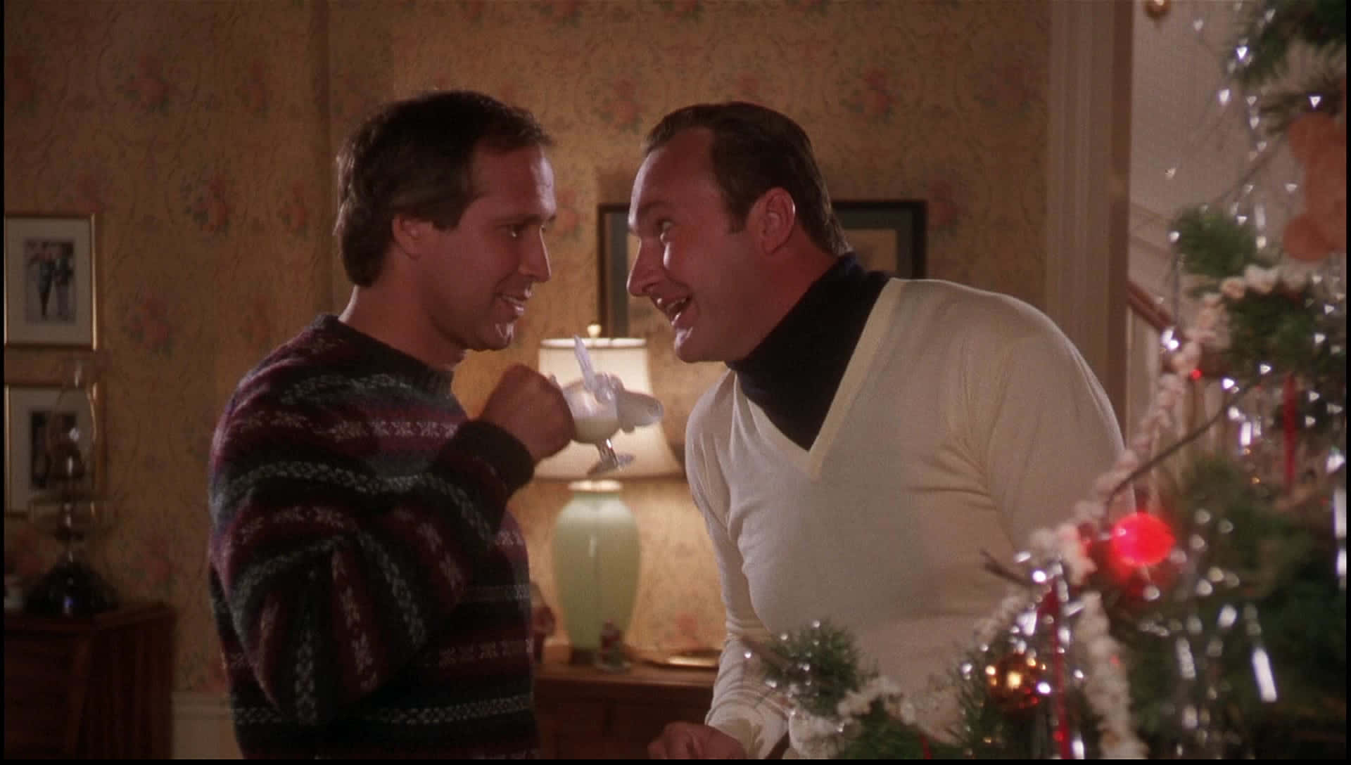 Christmas Vacation 桌布 壁紙 — 兩個男人在溫暖照明的客廳裡，在裝飾好的聖誕樹附近分享一個俏皮的時刻。