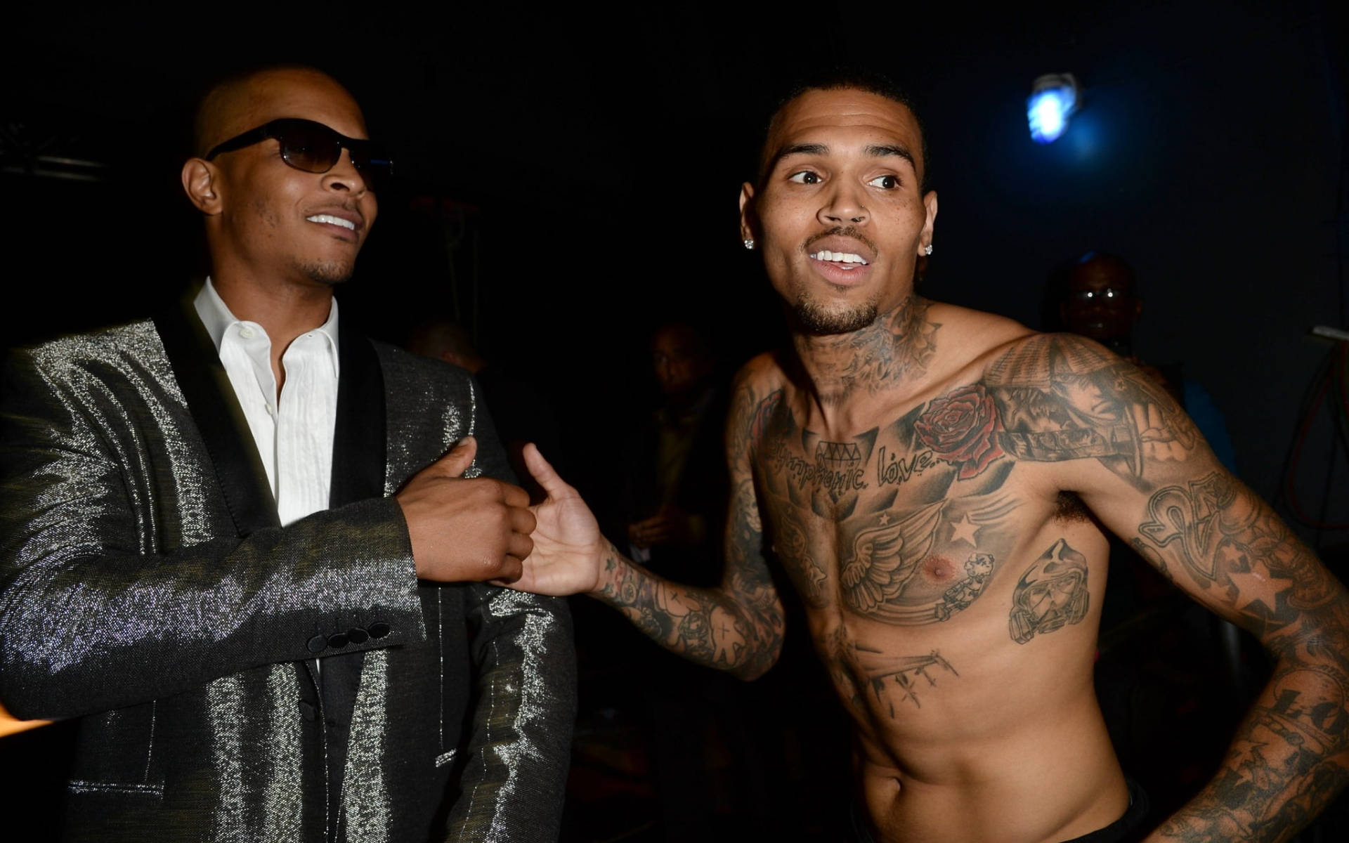 Chris Brown 桌布 壁紙 — 兩個男人在燈光昏暗的活動中自信地擺姿勢，一個穿著閃亮外套，另一個有刺青且赤裸上身。