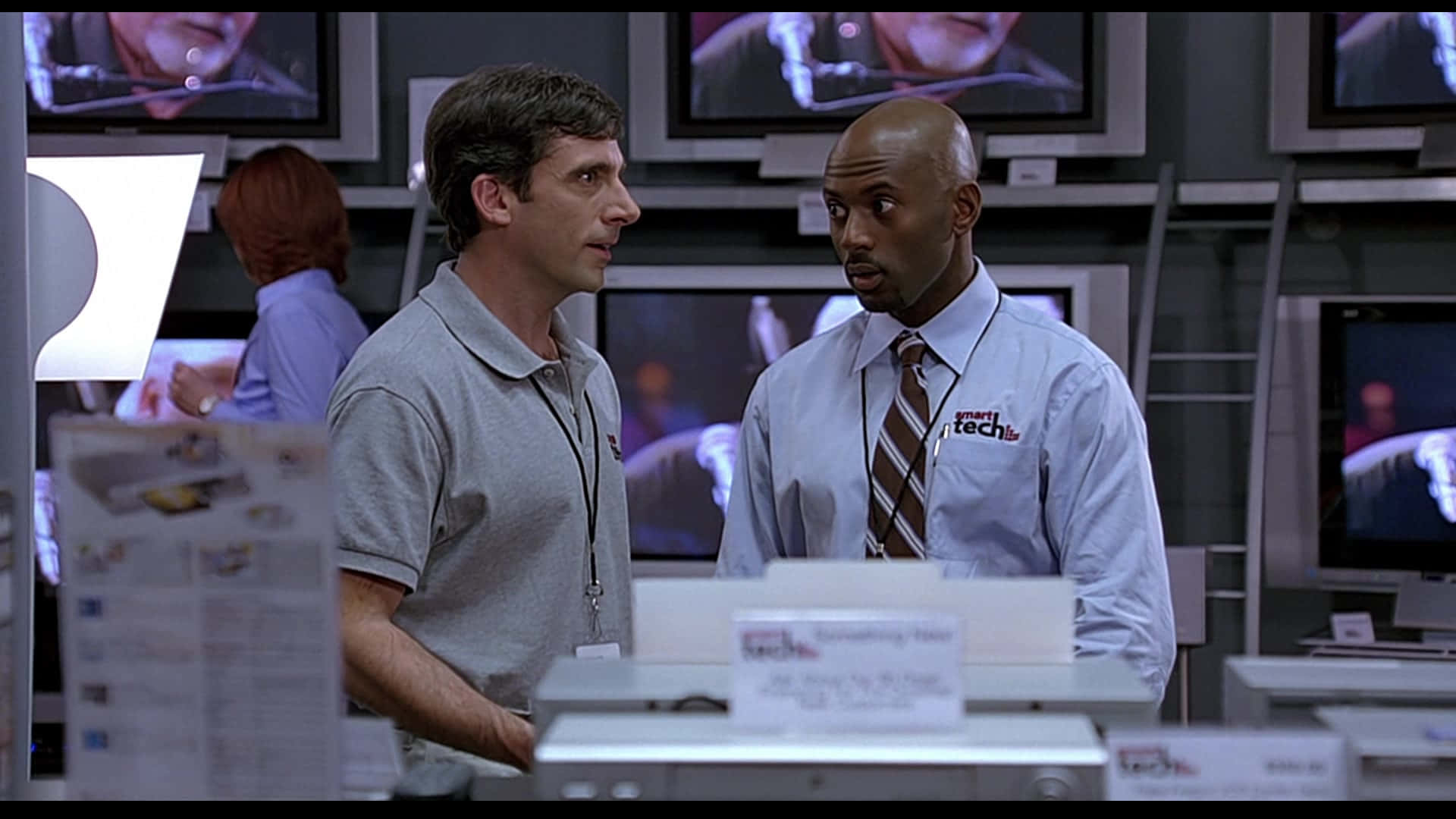 steve carell 桌布 壁紙 — 兩個男人在科技店裡，在電子產品貨架和發光螢幕之間進行嚴肅對話。
