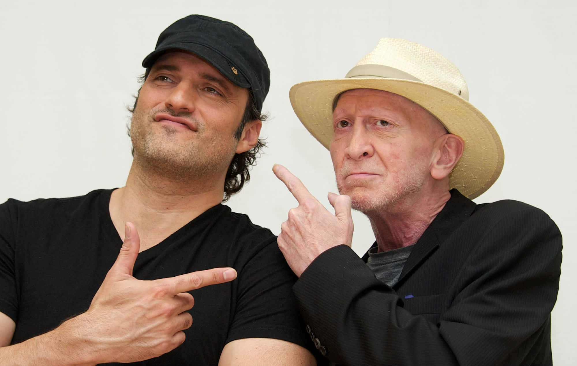 Frank Miller 桌布 壁紙 — 兩個男人在純白色背景前擺出俏皮姿勢，一個戴黑色鴨舌帽指著另一個戴草帽的男人，對方也指回去。