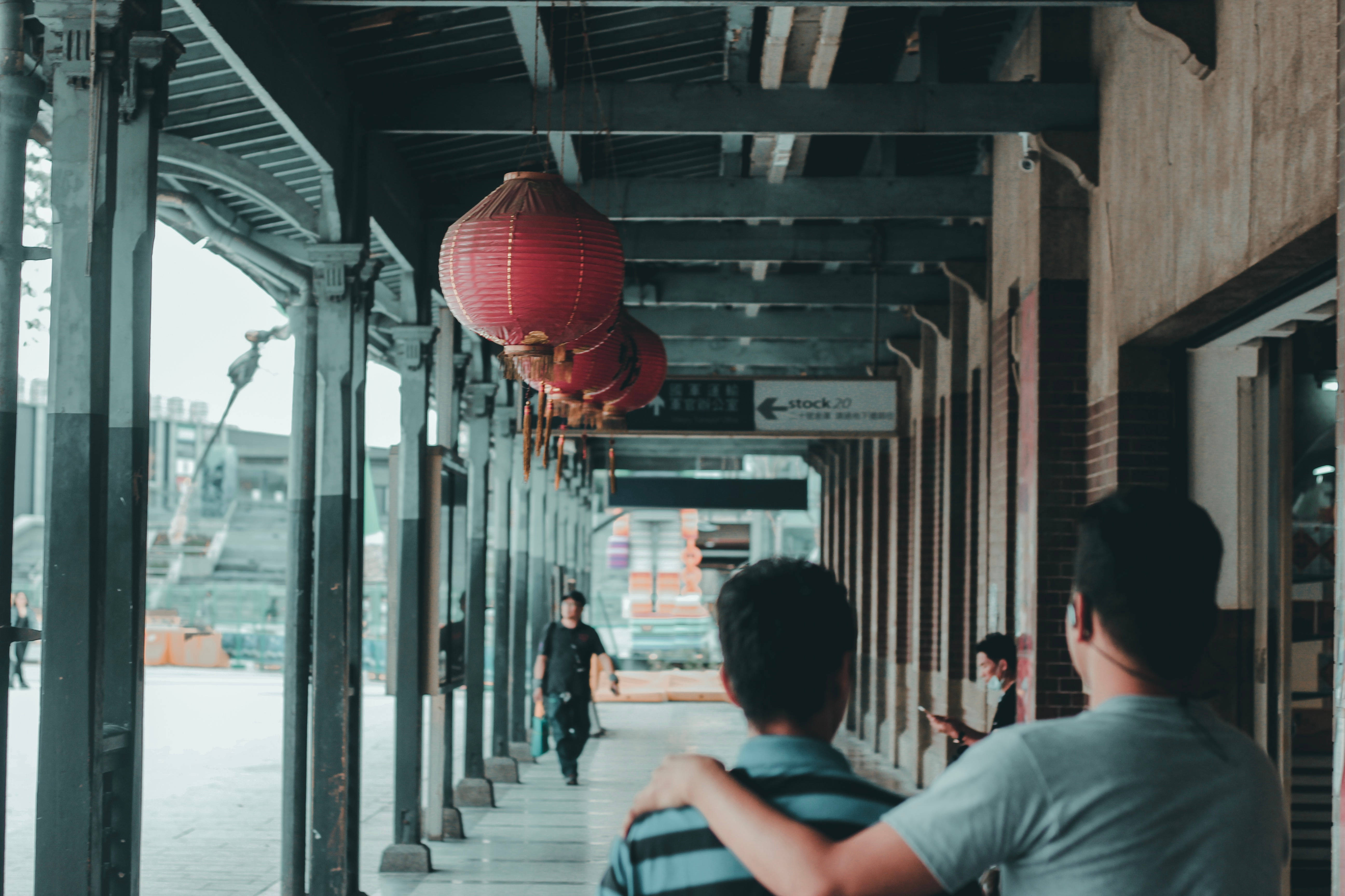 勵志動圖 桌布 壁紙 — 兩個男人在繁華的城市小巷中，走在一條有紅色燈籠懸掛在頭頂的有蓋行人道上。