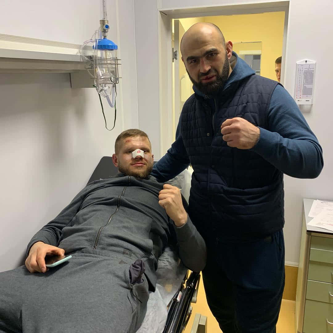 MMA 桌布 壁紙 — 兩個男人在醫院病房，一個坐在床上，帶著鼻導管和包紮的鼻子，另一個站在他旁邊擺出戰鬥姿勢。