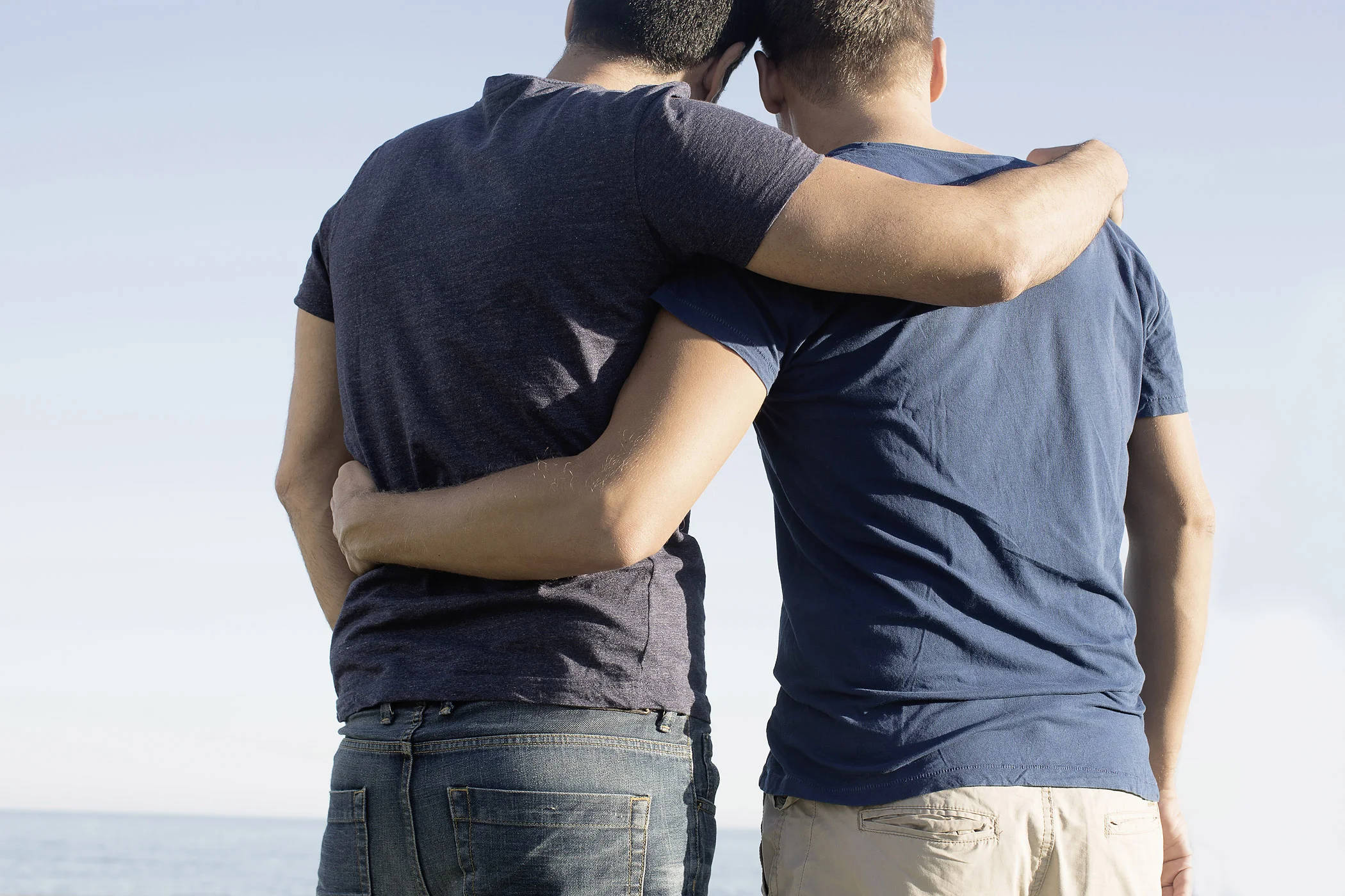 gay hug 桌布 壁紙 — 兩個男人從背後擁抱，一起站在晴朗天空下平靜的海洋前。