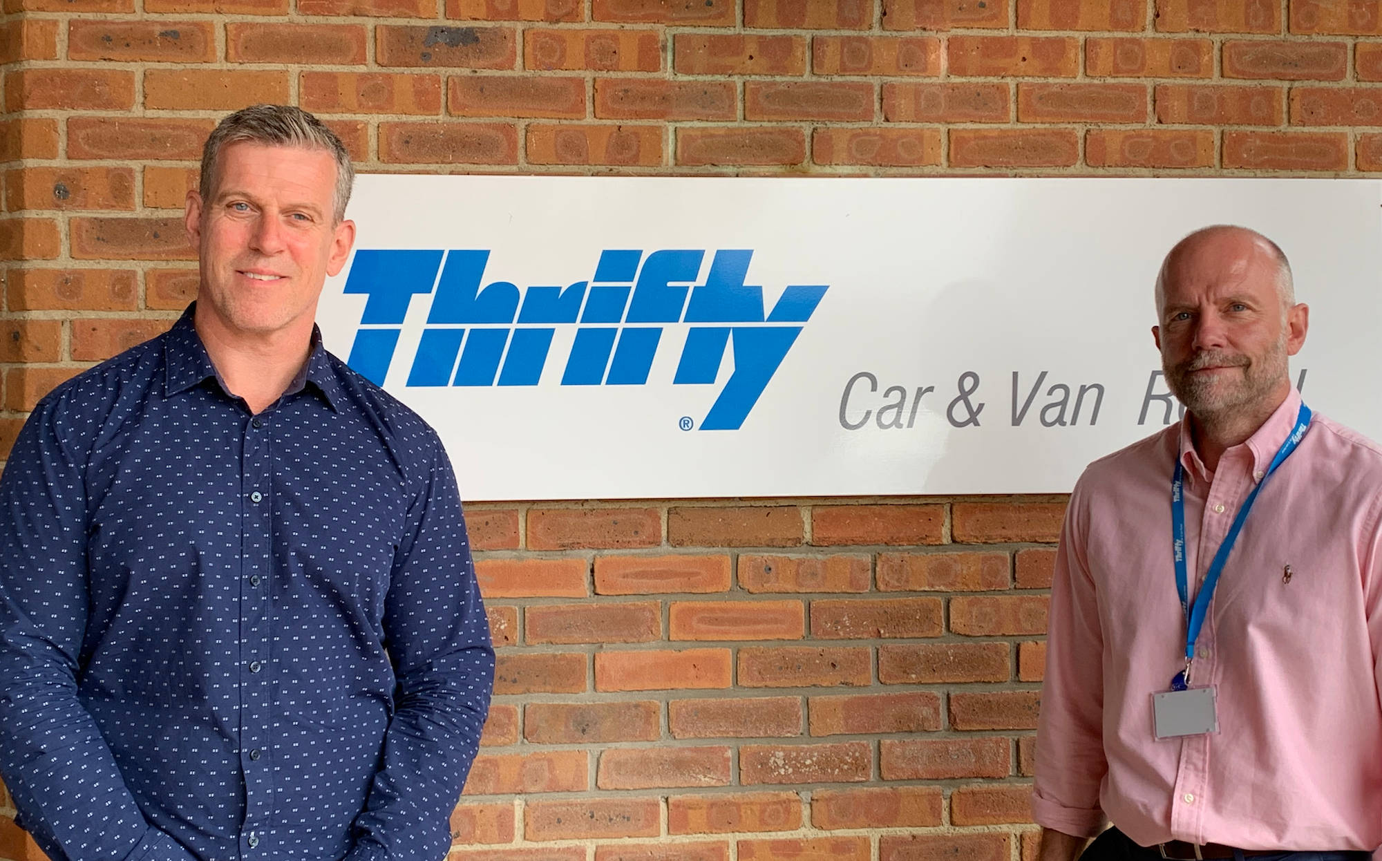 商業 桌布 壁紙 — 兩個男人微笑著站在磚牆前，牆上有 Thrifty Car & Van Rental 標誌。