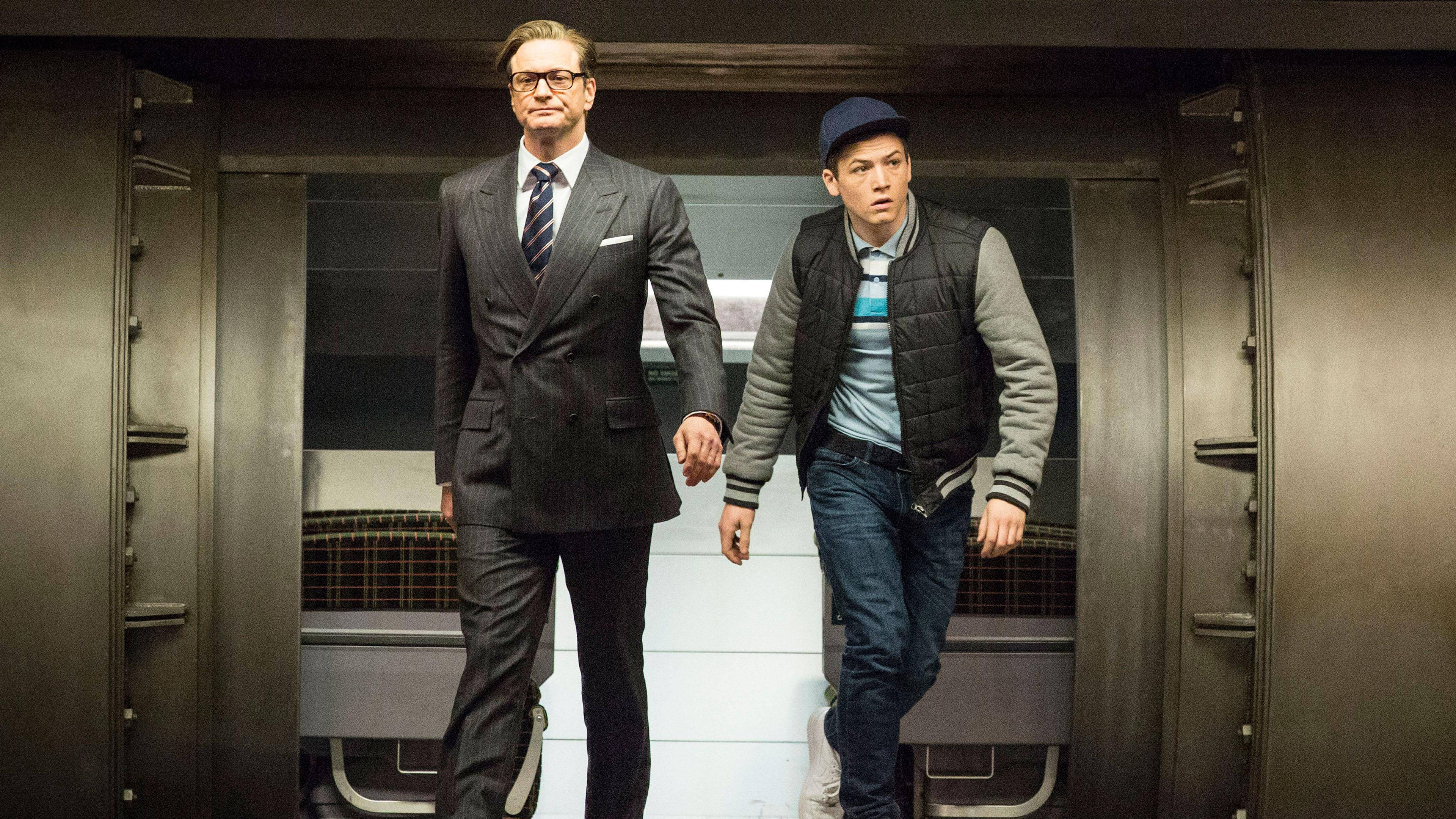 Kingsman 桌布 壁紙 — 兩個男人手牽手穿過金屬走廊，一個穿西裝，另一個穿背心和帽子。