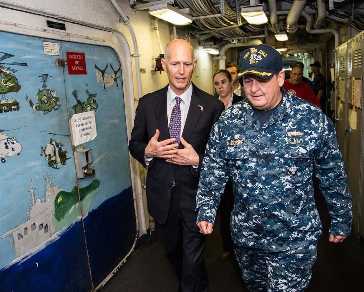 US Navy 桌布 壁紙 — 兩個男人穿過船艙走廊，一個穿西裝，另一個穿海軍制服，經過一扇繪有直升機和船隻的彩繪門。