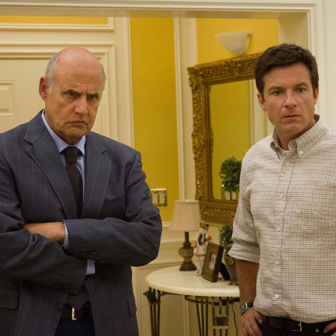 jeffrey tambor 桌布 壁紙 — 兩個男人站在一個光線溫暖、黃色牆壁的房間裡，一個穿著西裝雙臂交叉，另一個穿著鈕扣襯衫，兩人都表情嚴肅。