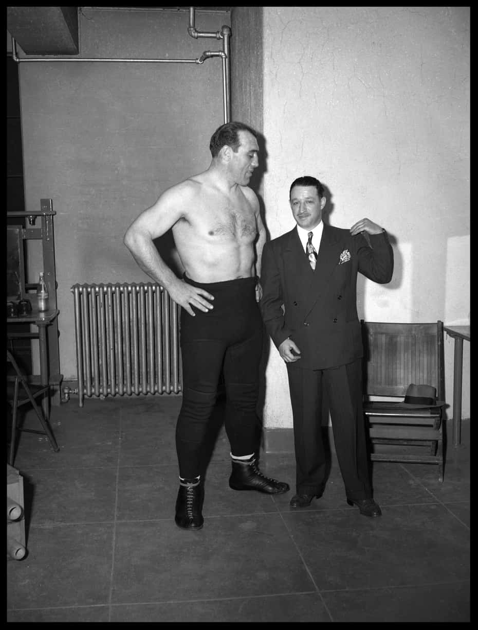 Boxing 桌布 壁紙 — 兩個男人站在一個空蕩的房間裡，一個赤裸上身穿著摔角緊身褲，另一個穿著西裝，手放在他的肩膀上。