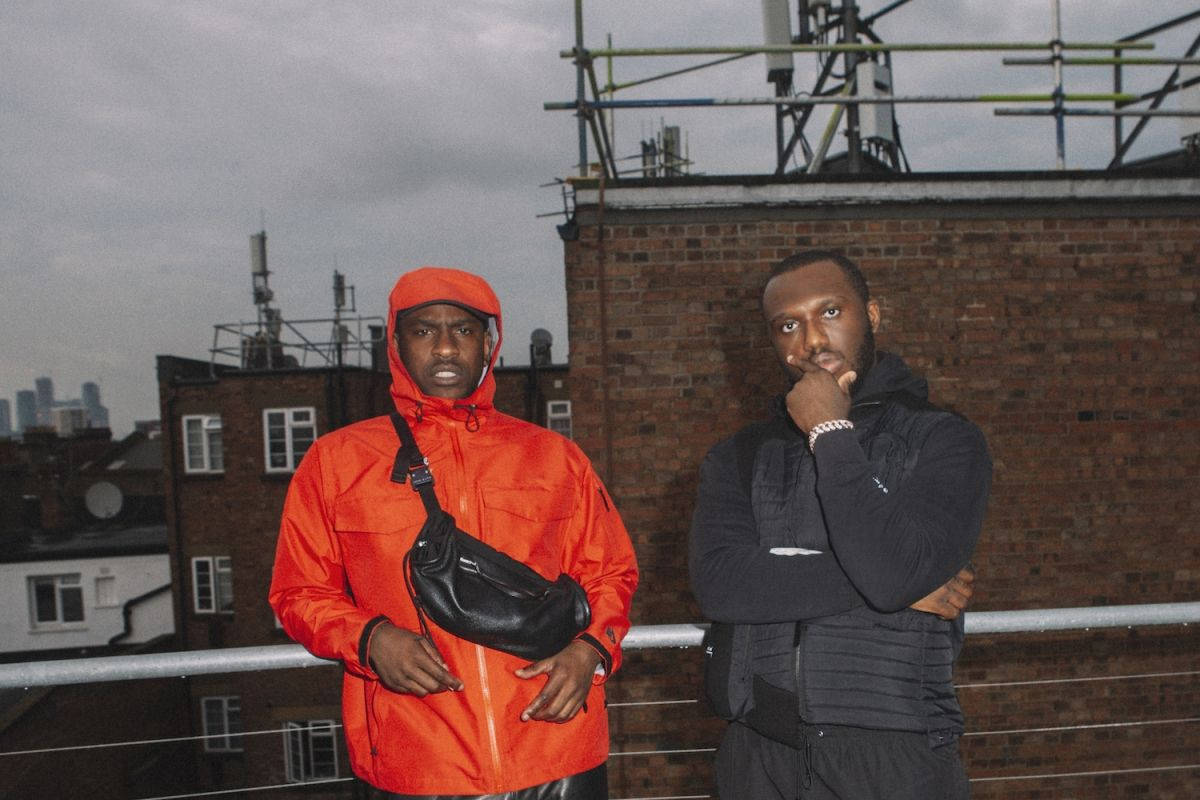 Skepta 桌布 壁紙 — 兩個男人站在粗糙的城市屋頂上，一個穿著亮橙色外套，另一個穿著深色裝備，背景是陰天。