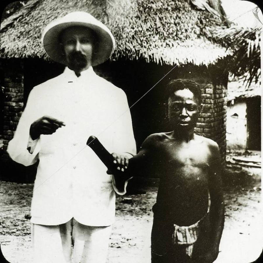 Congo 桌布 壁紙 — 兩個男人站在茅草屋前，一個穿白色西裝戴帽，另一個赤膊拿著瓶子。