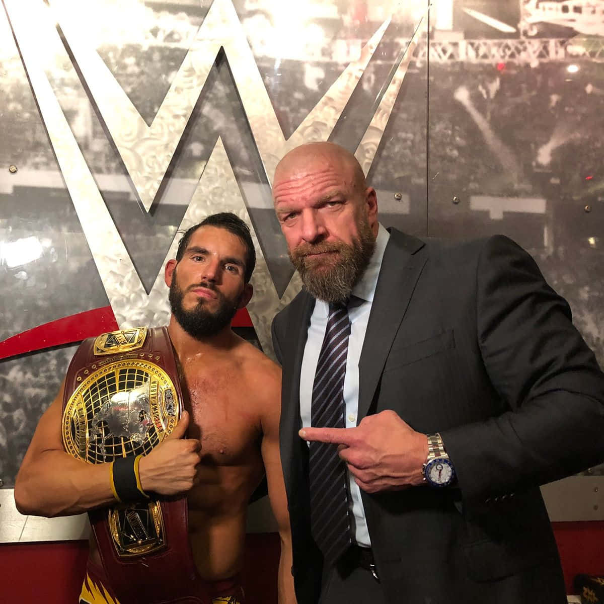 WWE 桌布 壁紙 — 兩個男人自信地擺姿勢，一個赤裸上身拿著冠軍腰帶，另一個穿著西裝指著他，背景是WWE標誌。