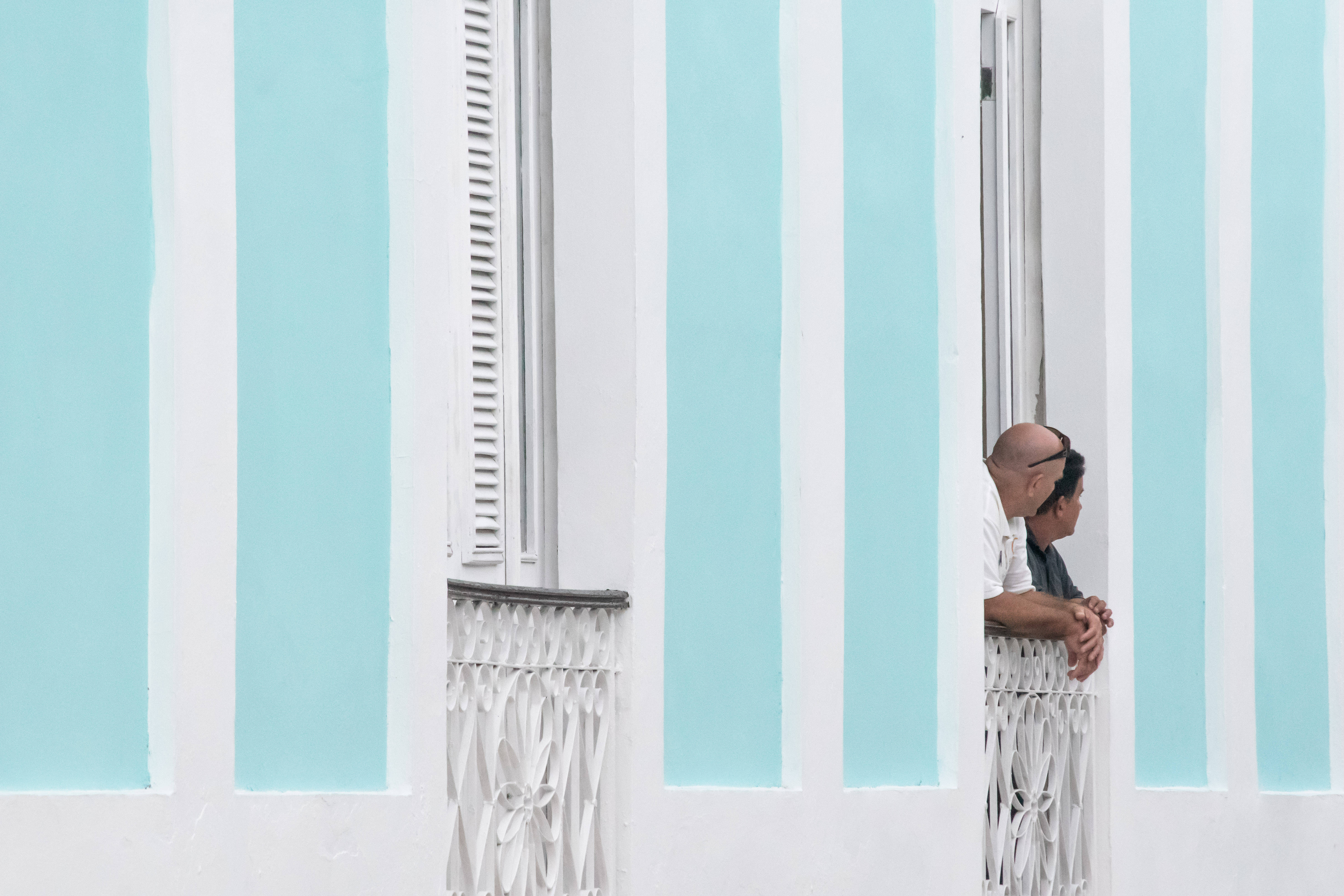 Cuba 桌布 壁紙 — 兩個男人靠在白色陽台欄杆上，以粉藍色和白色條紋牆為背景，向外凝望。
