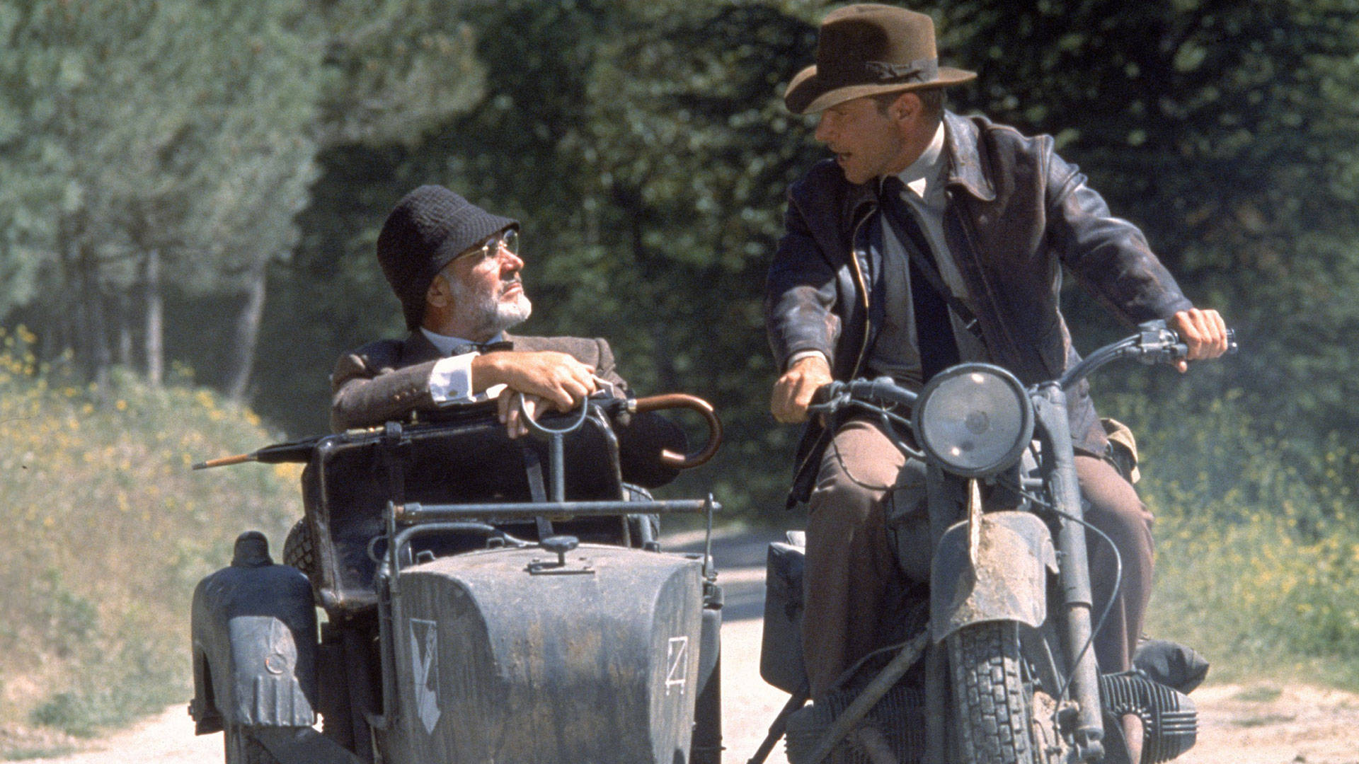 Indiana Jones 桌布 壁紙 — 兩個男人騎著一輛復古摩托車和側車，行駛在陽光普照、樹木和野花點綴的森林道路上。