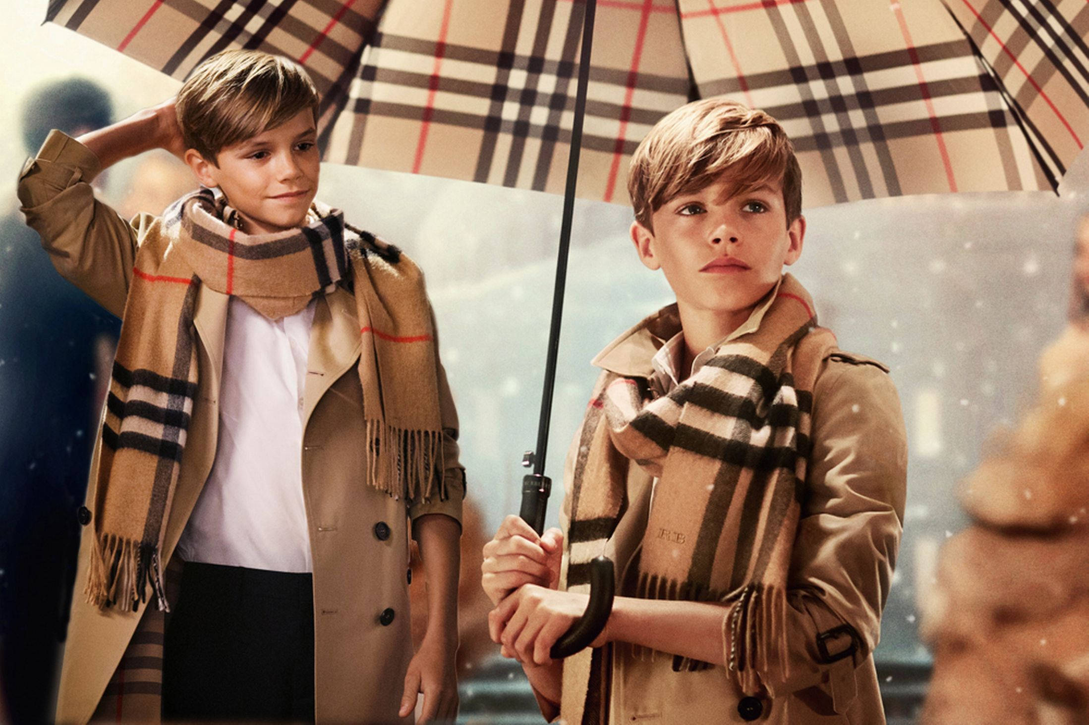 Burberry 桌布 壁紙 — 兩個男孩穿著米色風衣和相配的圍巾，站在格子雨傘下，雪花輕輕飄落在他們周圍。