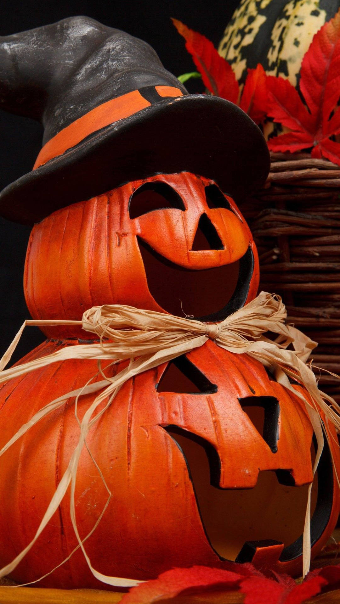 Halloween Phone 桌布 壁紙 — 兩個疊起的雕刻南瓜，戴著巫婆帽和稻草結，在秋葉和編織籃子的映襯下溫暖地發光。