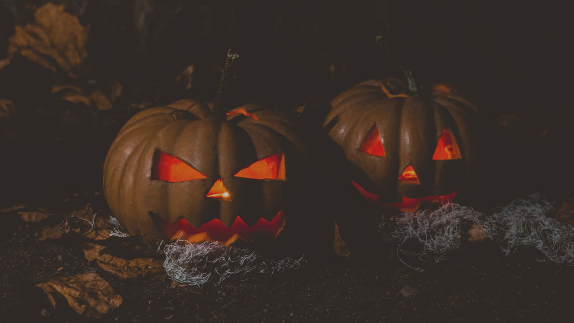 Halloween Pumpkin 桌布 壁紙 — 兩個發光的傑克南瓜燈，帶有尖銳的三角形眼睛和鋸齒狀牙齒，坐在昏暗光線下的落葉中。