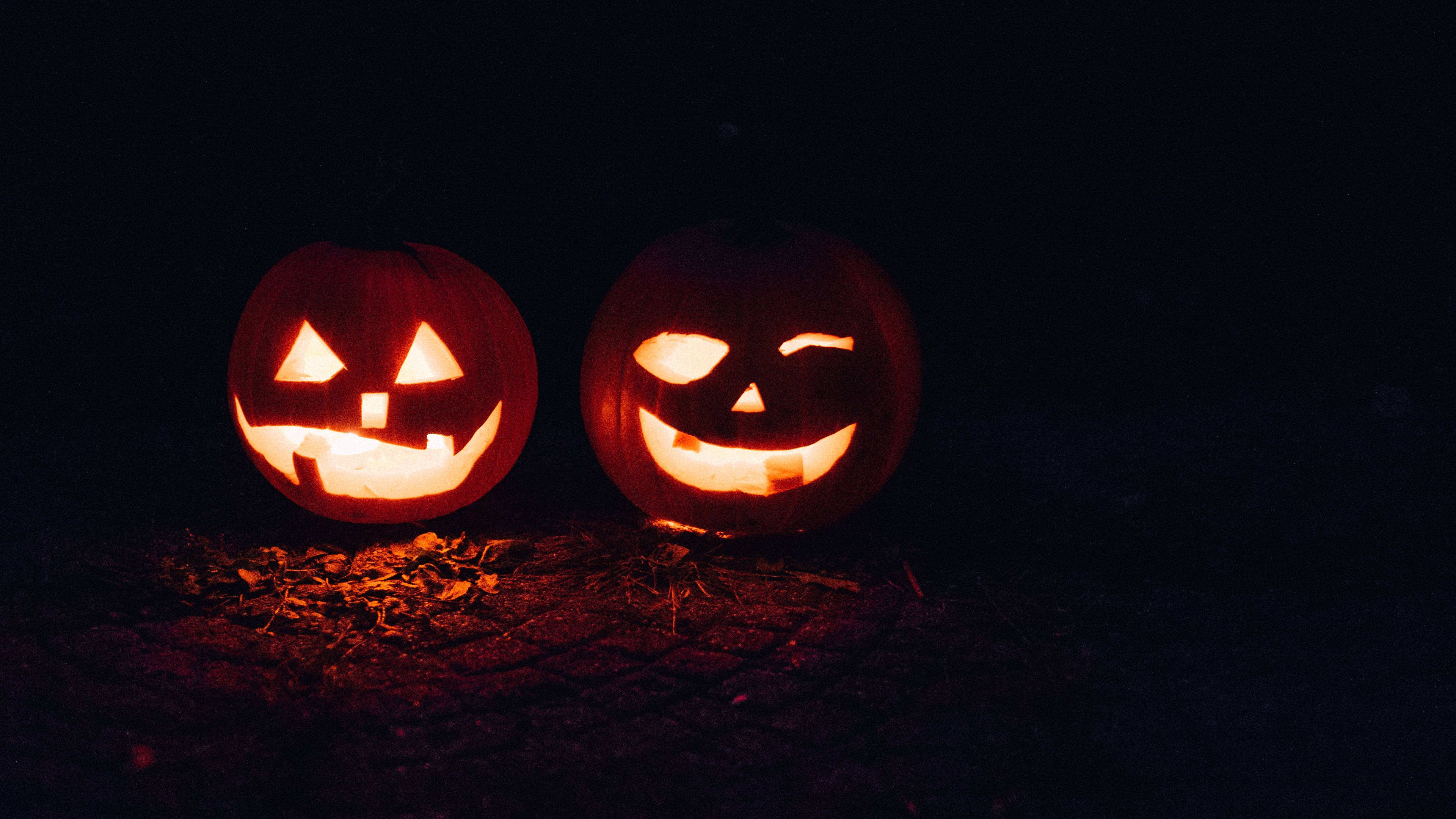 Cute October 桌布 壁紙 — 兩個發光的傑克南瓜燈，雕刻著臉孔，並排坐在黑暗的地面上，在夜晚散發出溫暖的橙色光芒。