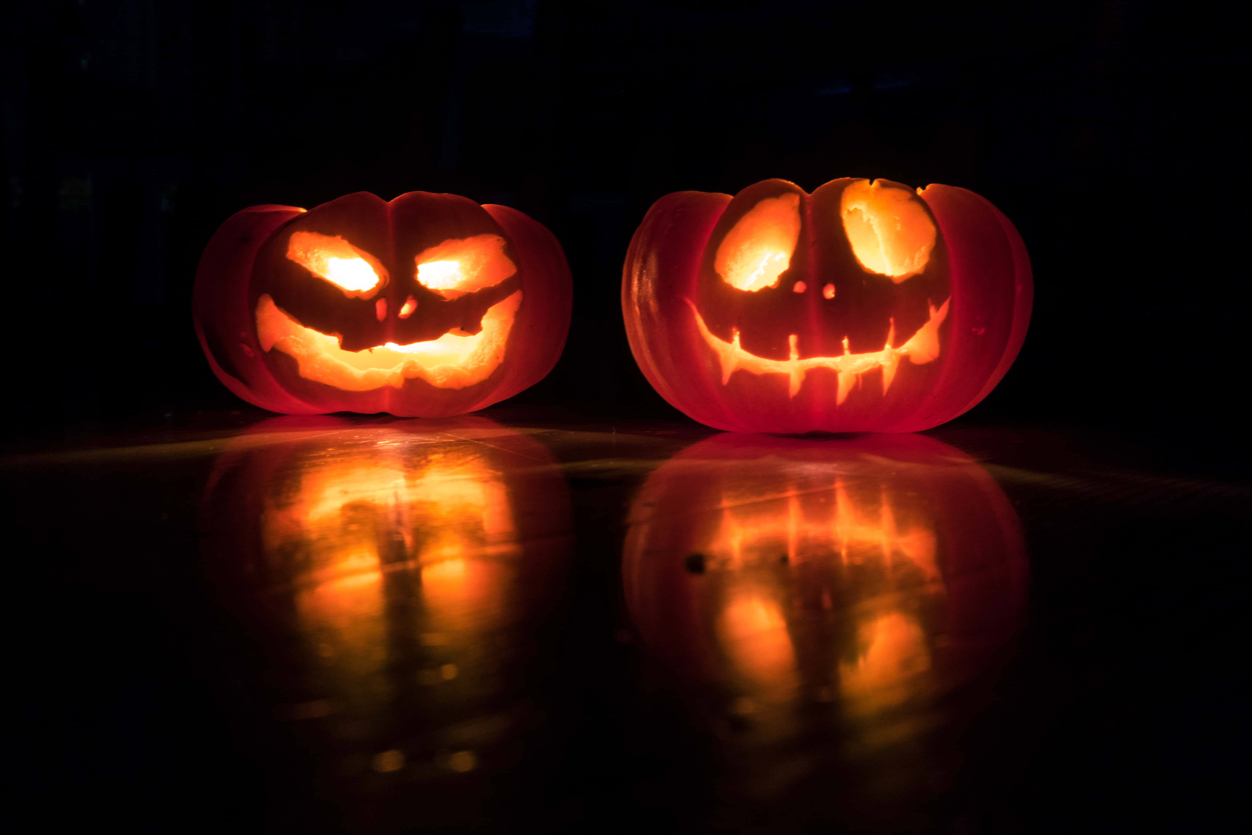 Halloween Macbook 桌布 壁紙 — 兩個發光的南瓜燈，帶有威脅性和咧嘴笑的面孔，在深色拋光表面上投射出詭異的倒影。