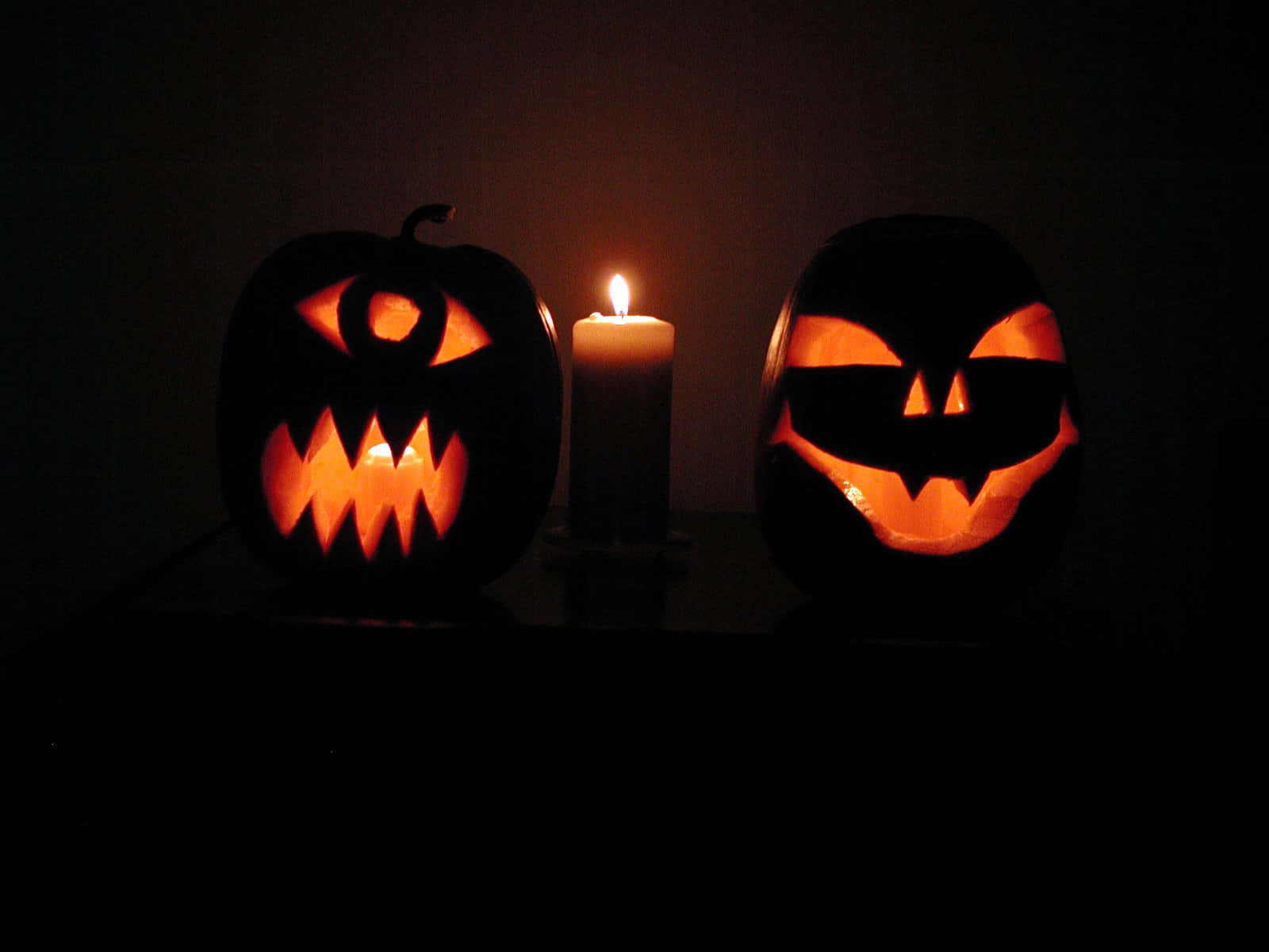 Jack O Lantern 桌布 壁紙 — 兩個發光的南瓜燈籠，帶有兇惡的面孔，在黑暗的房間中夾著一根閃爍的蠟燭。